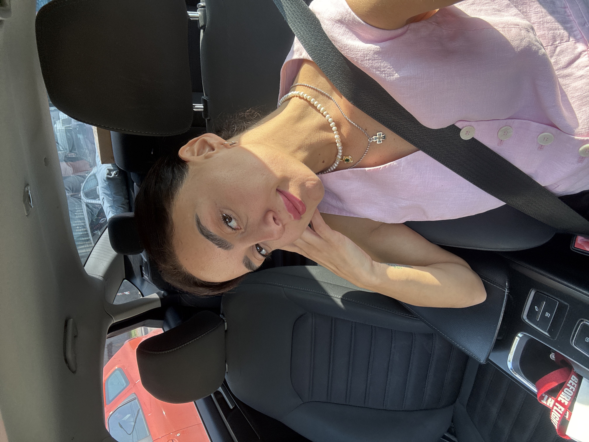 A woman is seated in the passenger seat of a car, looking at the camera. She is wearing a pink linen dress and a pearl necklace with a cross pendant. The car interior is visible, including the black leather seats and the center console. Outside the car, other vehicles are visible. The image appears to have been taken during the daytime, with sunlight casting shadows. The overall scene suggests a casual, everyday moment captured on the go.