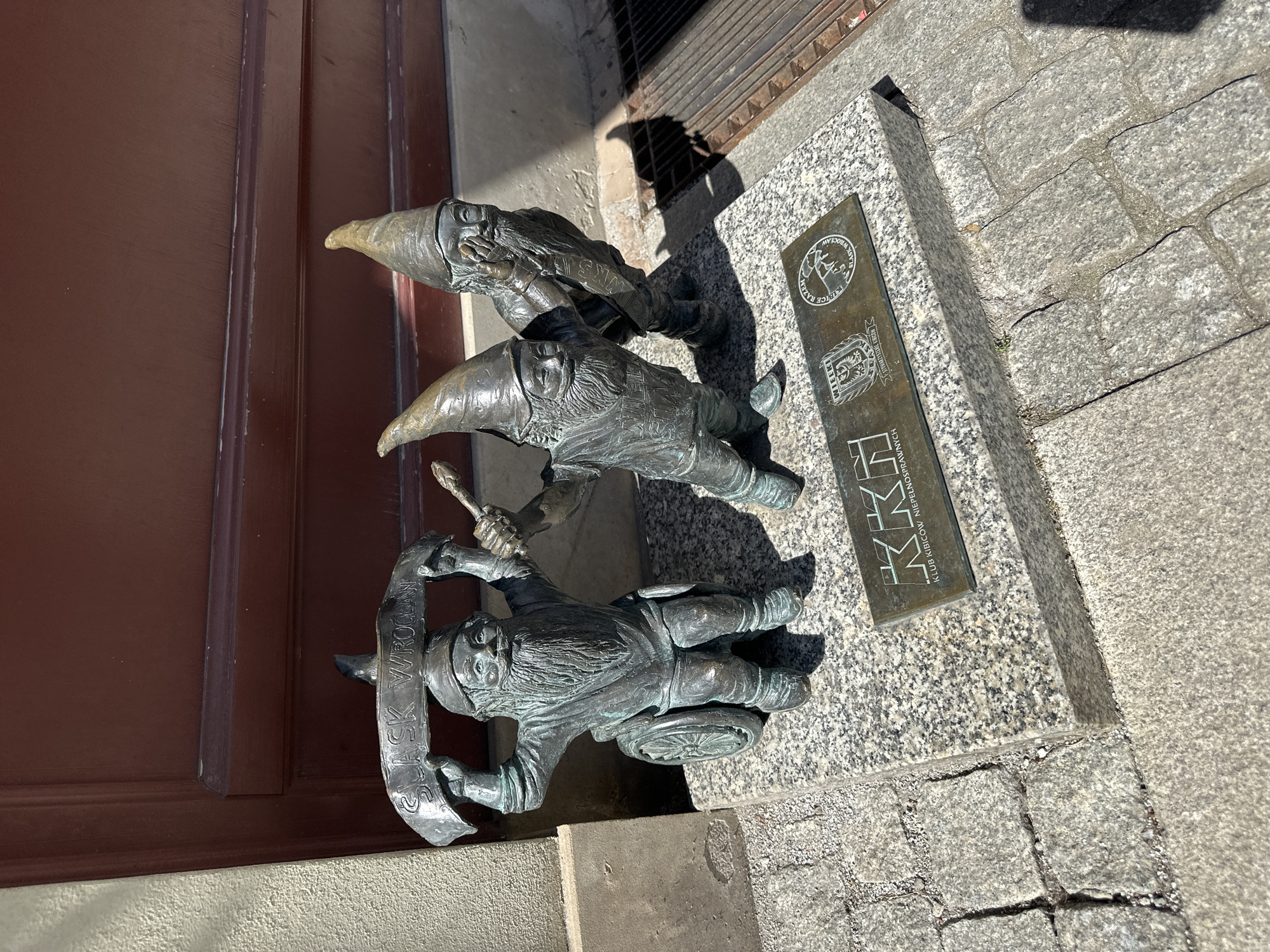 This is a photograph of a statue depicting three dwarves in Wrocław, Poland. The dwarves are made of bronze and are arranged in a line, one behind the other. The first dwarf is holding a banner that reads "SLASK WROCLAW". The second dwarf is holding a brush, and the third dwarf is covering his mouth with his hand. The statue is located on a stone platform next to a bronze plaque that reads "KKH KLUB KIBICOW NIEPELNOSPRAWNYCH". The statue is located next to a brown door. The background consists of stone pavement.