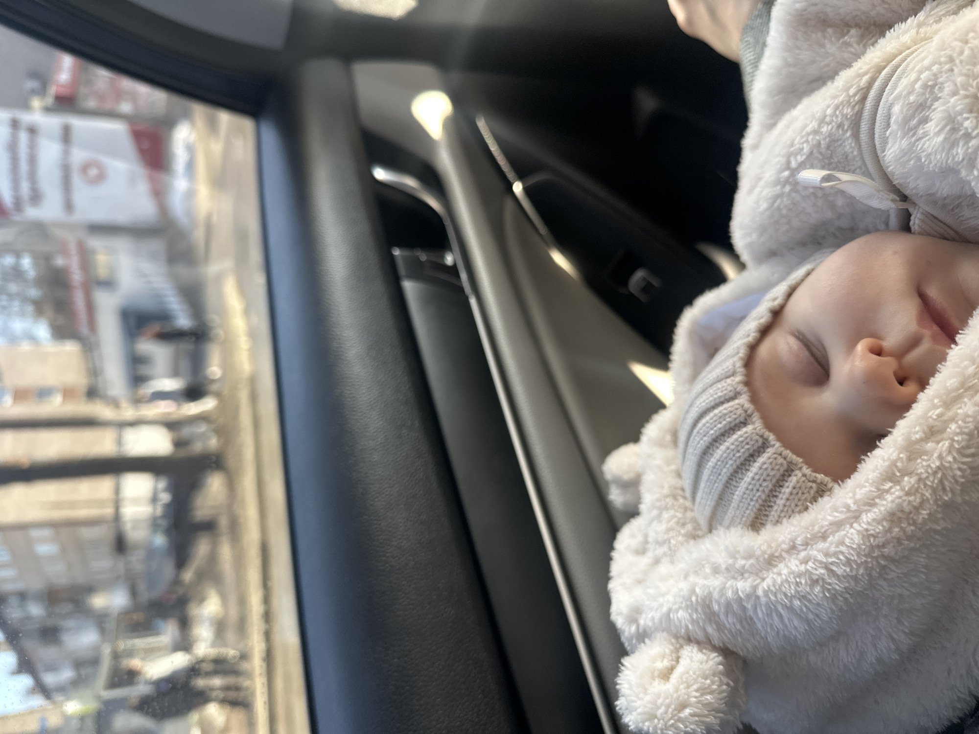 The image depicts a baby sleeping in a car seat inside a vehicle. The baby is wearing a white, fluffy hooded jacket over a grey knitted cap. The car's interior is visible, including the door panel and window. Outside the window is a blurry street scene with buildings, possibly located in Chaikovsky, Russia. The specific buildings or landmarks necessary to confirm the location are unclear from this image.