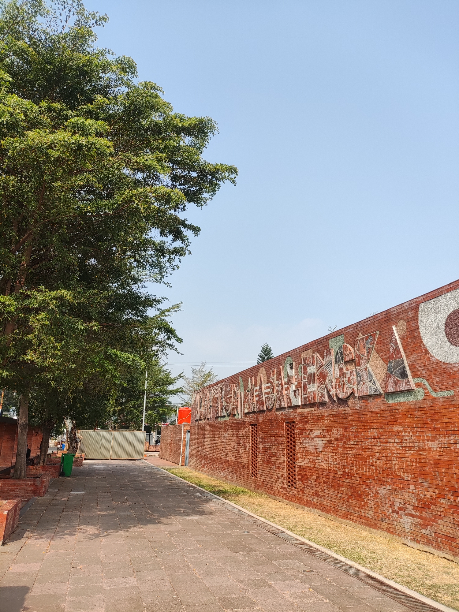The image captures a wide, paved pathway leading through an outdoor area. To the left, a large tree with dense green foliage shades part of the walkway. Brick planters and low brick walls line the edge of the pathway beneath the tree. Further down the path, a tall, corrugated metal fence is visible, with a green trash can placed near its base.
On the right side of the pathway, a prominent brick wall stretches into the distance. This wall is adorned with a mosaic mural that spells out words in large, decorative letters. The mosaic features a variety of colors and patterns within each letter, along with some abstract shapes. The wall itself is constructed of red bricks, with visible mortar lines. In the background, beyond the wall, the sky is a clear, pale blue. The overall environment appears to be a public outdoor space, possibly a park or a pedestrian area within a city. The scene is well-lit by natural sunlight, casting shadows on the pathway. The specific location in the city of Majalengka, Indonesia, is not visually indicated by any landmarks or signage in the provided image.