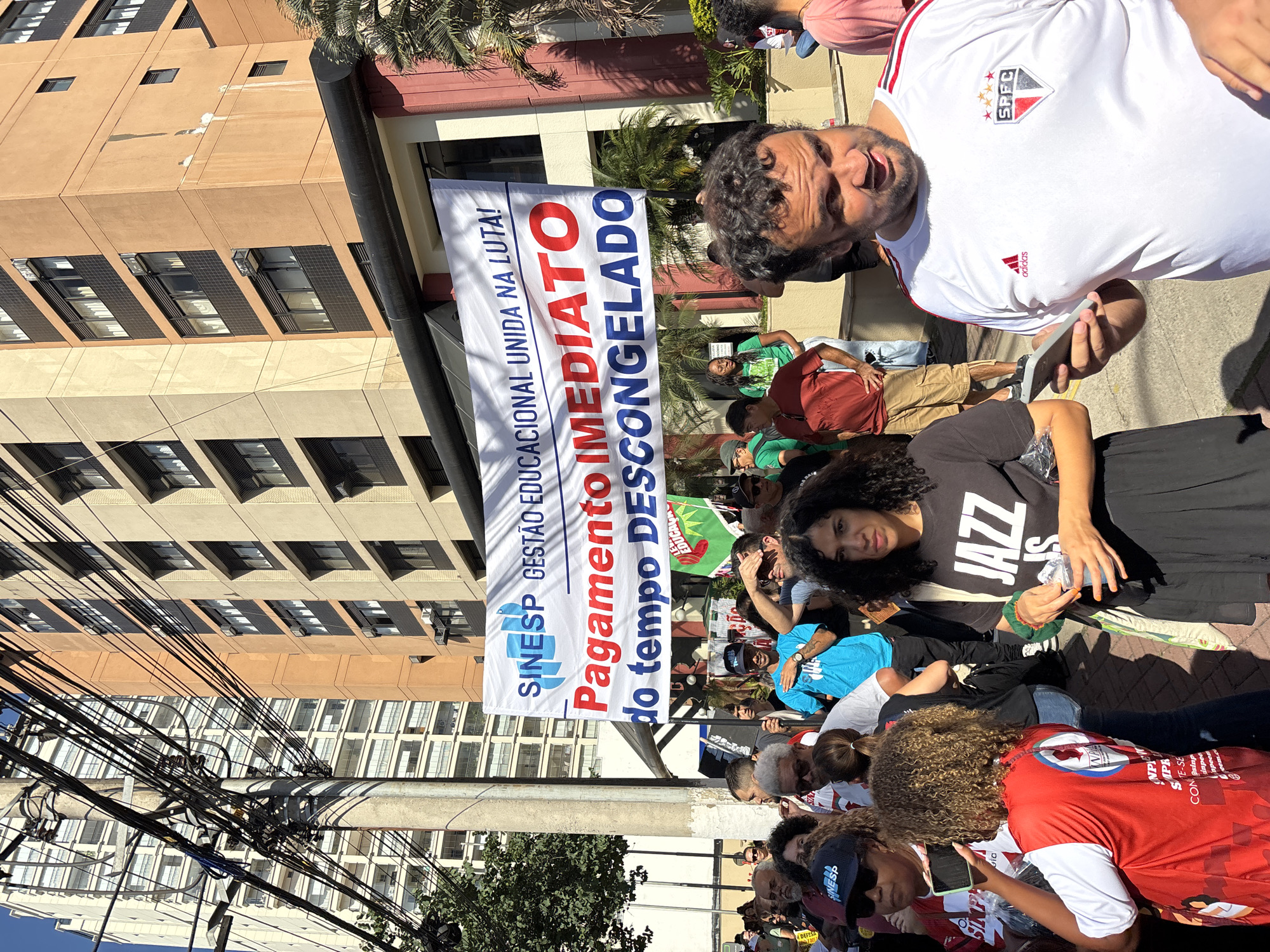 A crowd of people is gathered on a street in what appears to be a protest or demonstration. A large banner hangs behind them, featuring the text "SINESP GESTÃO EDUCACIONAL UNIDA NA LUTA! Pagamento IMEDIATO do tempo DESCONGELADO" in Portuguese, suggesting the event is related to education and demanding immediate payment. A tall building stands in the background, indicating an urban setting, most likely in the city of São Paulo, Brazil. Power lines crisscross overhead. People are dressed in various attire, with some wearing shirts with the "SPFC" logo, indicating support for the São Paulo Futebol Clube. Some individuals are holding phones, possibly recording or taking photos of the event. The environment suggests a sunny day, with shadows cast on the ground.