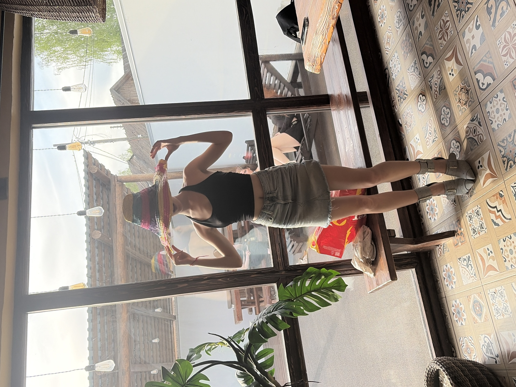 A woman stands in front of a large multi-pane window at a restaurant in Старое Михайловское, Russia. She is wearing a black tank top, a beige skirt, and grey buckled boots, and she is holding a rainbow sombrero. The interior flooring is patterned tile. Outside the window, a building with hanging lightbulbs can be seen. A green leafy plant is situated near the bottom of the window.