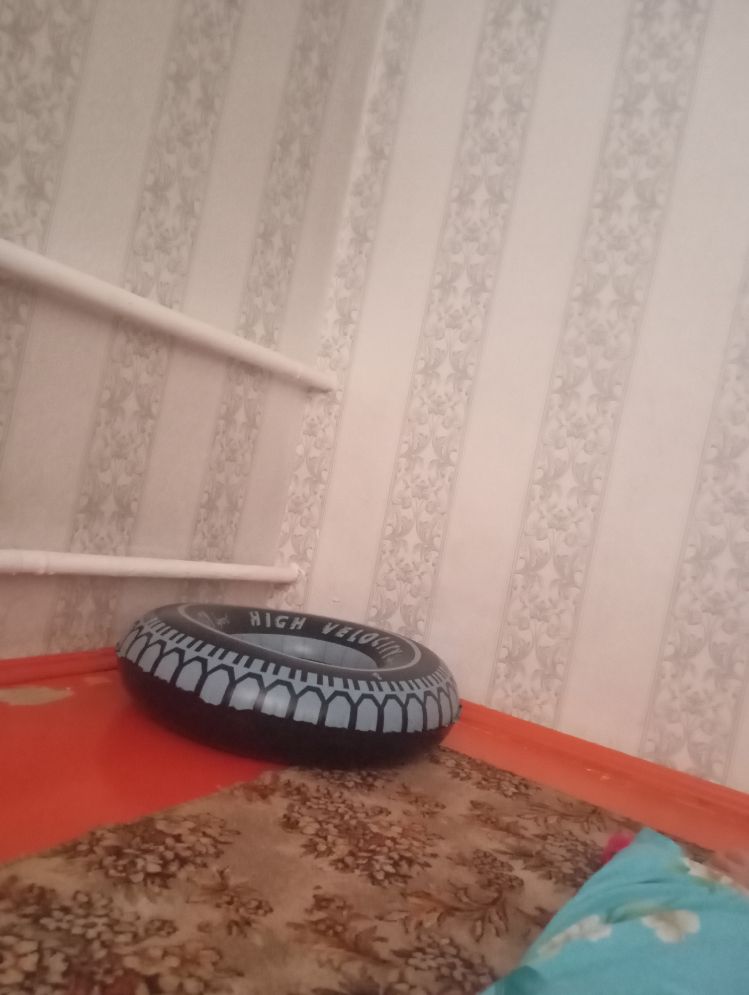 A partially inflated, black and blue inflatable ring with the words "HIGH VELOCITY" printed on it rests on an orange surface. Behind the ring, a patterned carpet is visible, and to the right of the ring, a light blue pillow with a floral pattern is partially in view. The background consists of a wall with striped wallpaper featuring a repeating floral motif. Two white pipes, likely radiators or conduits, are mounted horizontally on the wall. The scene appears to be indoors, possibly a living space in Arys, Kazakhstan. There are no people or animals present in the image.