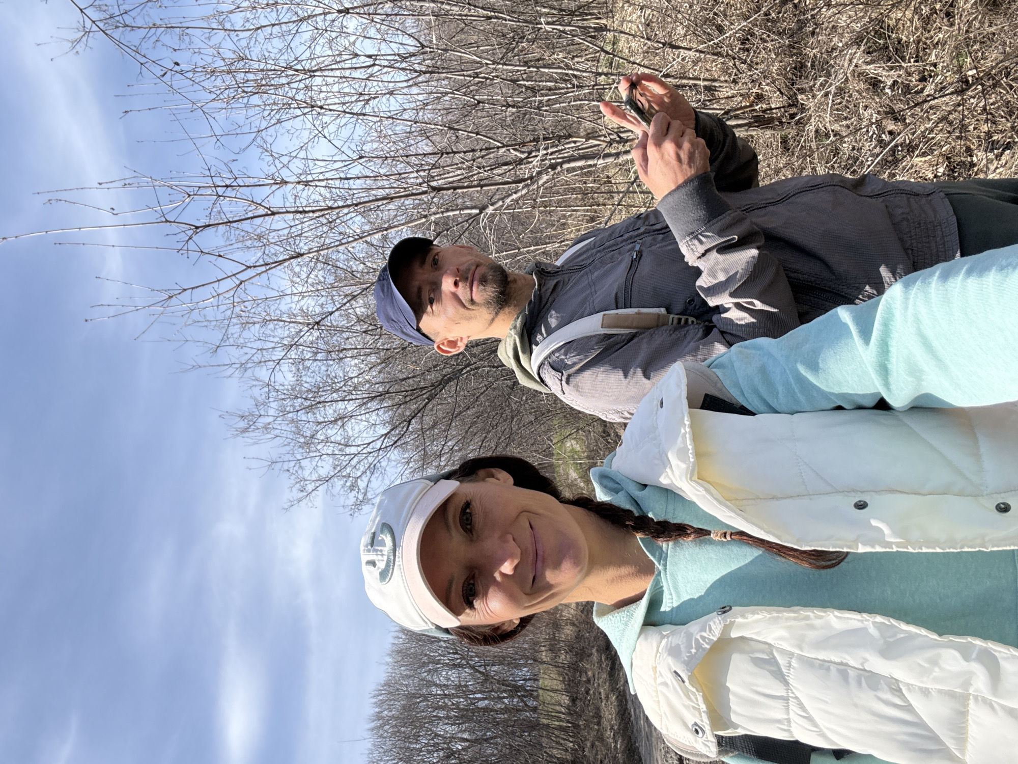 This media file captures two individuals outdoors in Ulyanovsk, Russia, against a backdrop of bare trees and a clear blue sky. The image is rotated, presenting the subjects as if lying down, but they are upright.
In the foreground on the left, a woman smiles faintly while looking directly at the camera. She wears a light-colored baseball cap with a prominent "G" logo on the front, a light blue hooded sweatshirt, and a white quilted puffer vest. Her dark hair is styled in a braid.
Behind her and to the right is a man with a beard, wearing a blue baseball cap, a grey zip-up jacket over a greenish hoodie, and dark pants. He is holding a smartphone with both hands, looking down at its screen. A light-colored strap or harness is visible across his chest.
The scene depicts a natural environment, likely a park or forest edge, characterized by numerous leafless trees and bushes, suggesting a late winter or early spring season. The ground is covered with dry, light brown undergrowth. The sky is bright blue with scattered thin clouds, indicating a clear, sunny day. Both individuals appear relaxed, seemingly enjoying an outdoor outing.