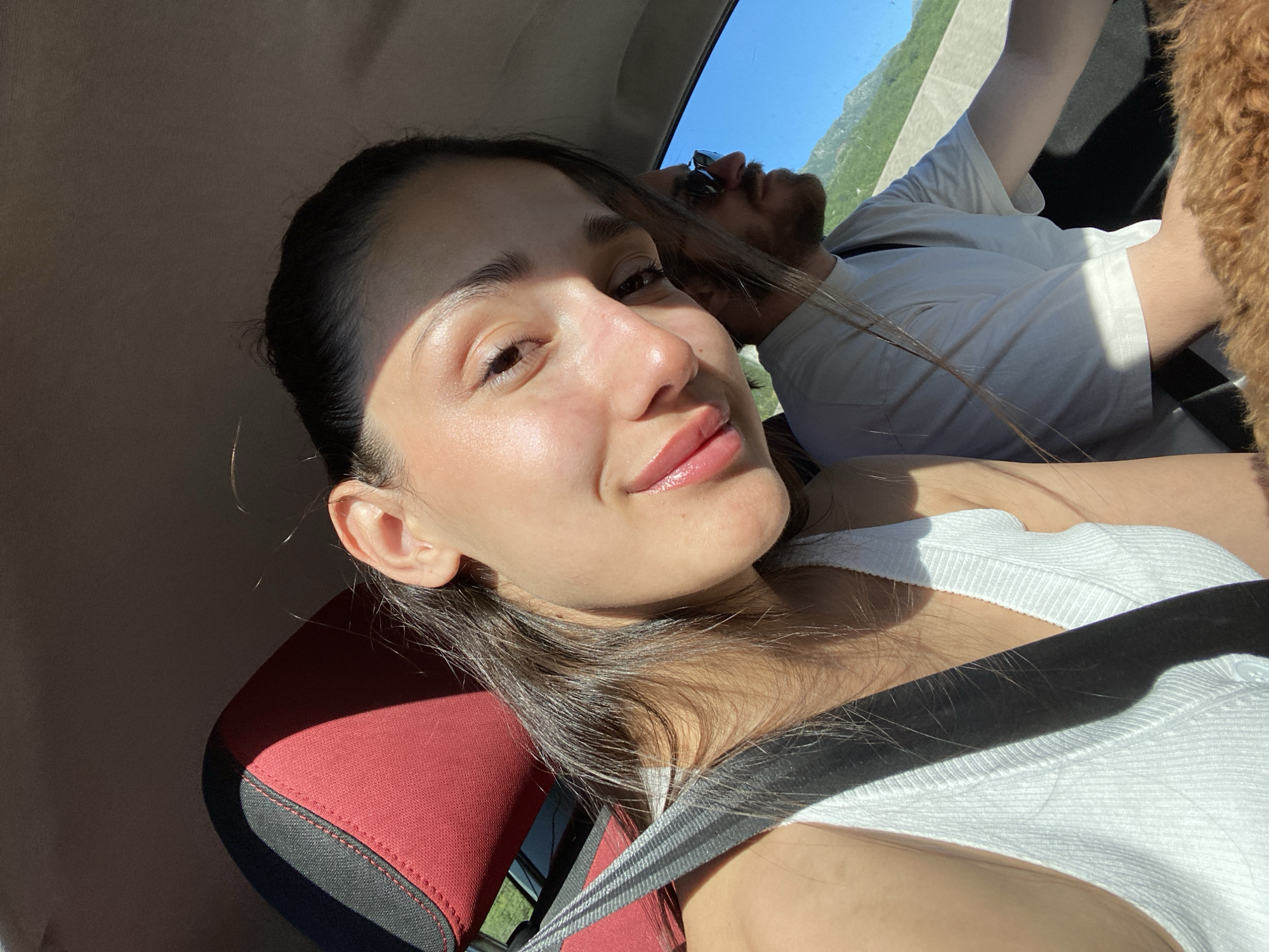 A woman is taking a selfie in a car. She has dark hair pulled back, wearing a white tank top and a seatbelt, and is looking at the camera with a slight smile. In the background, a man with sunglasses is visible, appearing to be asleep, wearing a white t-shirt. The car is moving, and sunlight is streaming in, with a glimpse of a mountainous landscape visible through the car window. The location appears to be Sotonići, Montenegro based on the landscape.