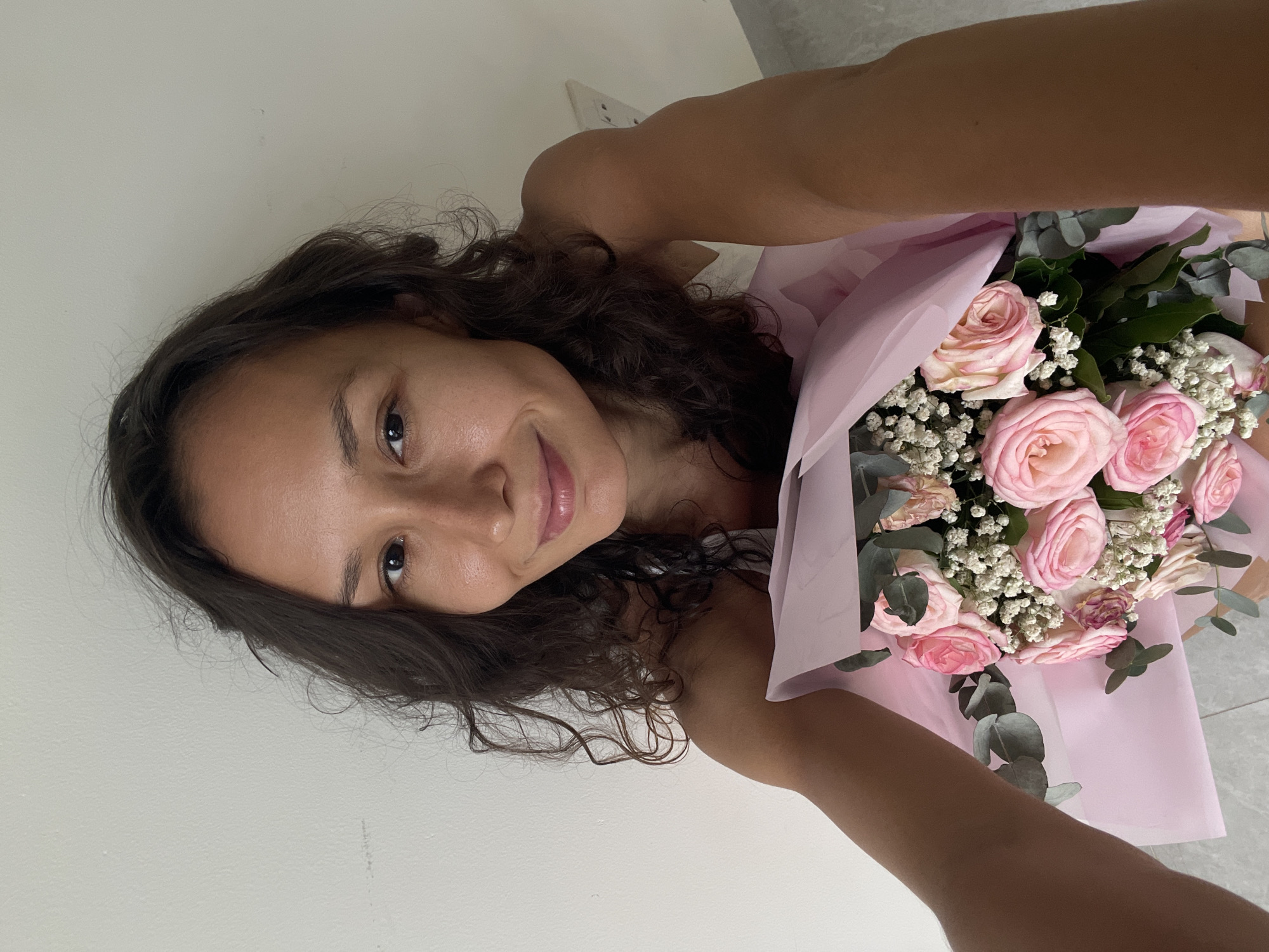 A woman with dark, curly hair, holding a bouquet of pink roses and baby's breath, is pictured from a slightly high angle. She is looking directly at the camera with a subtle smile. The background consists of a plain white wall and a tiled floor, suggesting an indoor setting. The bouquet is wrapped in pink paper. The image does not contain any elements that would indicate a specific location in Mui Ne, Vietnam.