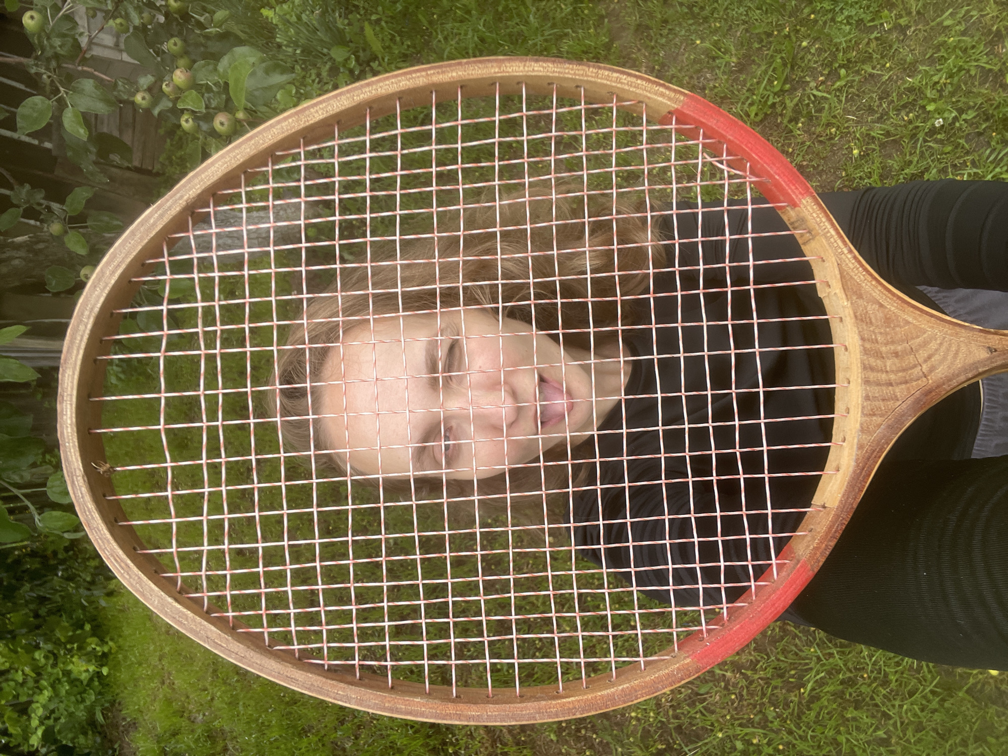 A person is lying on grass, viewed from above through the strings of a wooden tennis racket. The person's face is in the center of the frame, with their tongue sticking out. Their eyes are closed and they have light-colored hair. They are wearing a black top and dark pants. The tennis racket has a wooden frame and pink and white strings. A small branch with green apples is visible in the upper left corner, indicating an outdoor setting with foliage and grass. The scene appears to be set during the daytime, with natural lighting. The overall mood is playful and casual. No text is visible in the image.