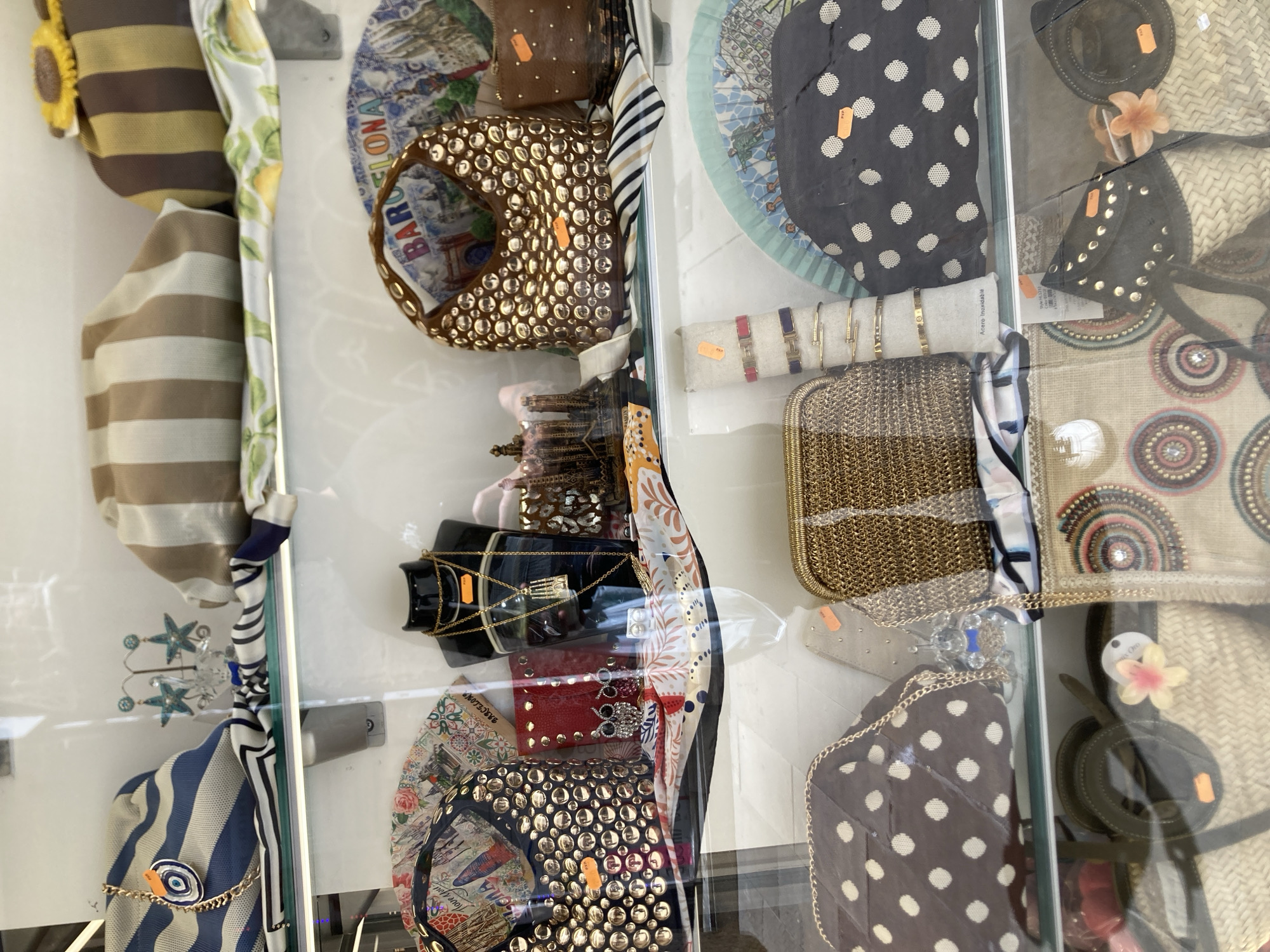 The image shows a close-up, high-angle view of a retail display case, possibly in a souvenir shop, exhibiting various accessories. The display is divided into vertical sections by glass.
Prominently featured are several purses and bags. One bag is a round, metallic gold purse adorned with numerous circular embellishments. Another is a smaller brown leather pouch with gold studs. Several scarves with various patterns, including stripes and floral designs, are also visible, some draped and others folded. There are also pieces of jewelry, including bracelets, displayed on a white stand. Some items have small orange tags attached, likely price tags.
In one section, a circular fan-like object with a map-like design and text suggesting "BARCELONA" is visible behind a studded purse. Another similar fan-like item with a dark background and white polka dots is also displayed.
Other items include a woven straw bag with decorative circles and a smaller, dark-colored hat with a flower embellishment. The overall impression is of a curated collection of fashion accessories and souvenirs. The lighting suggests an indoor setting, and the presence of price tags indicates it is a commercial establishment. The location context is identified as Barcelona, Spain, which aligns with the visible text on the fan-like item.