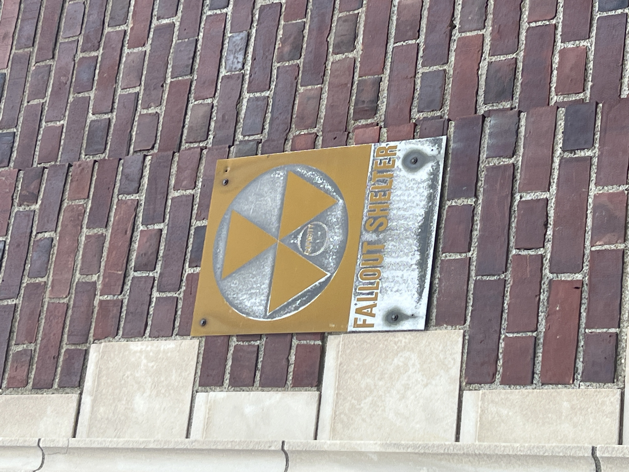 A Fallout Shelter sign is affixed to a brick wall in the City of Newburgh, United States of America. The sign features a radiation trefoil symbol within a circle. The sign reads "Fallout Shelter". The sign is attached to the wall with bolts. The brick wall is composed of red bricks. The sign is located above a light-colored stone wall.