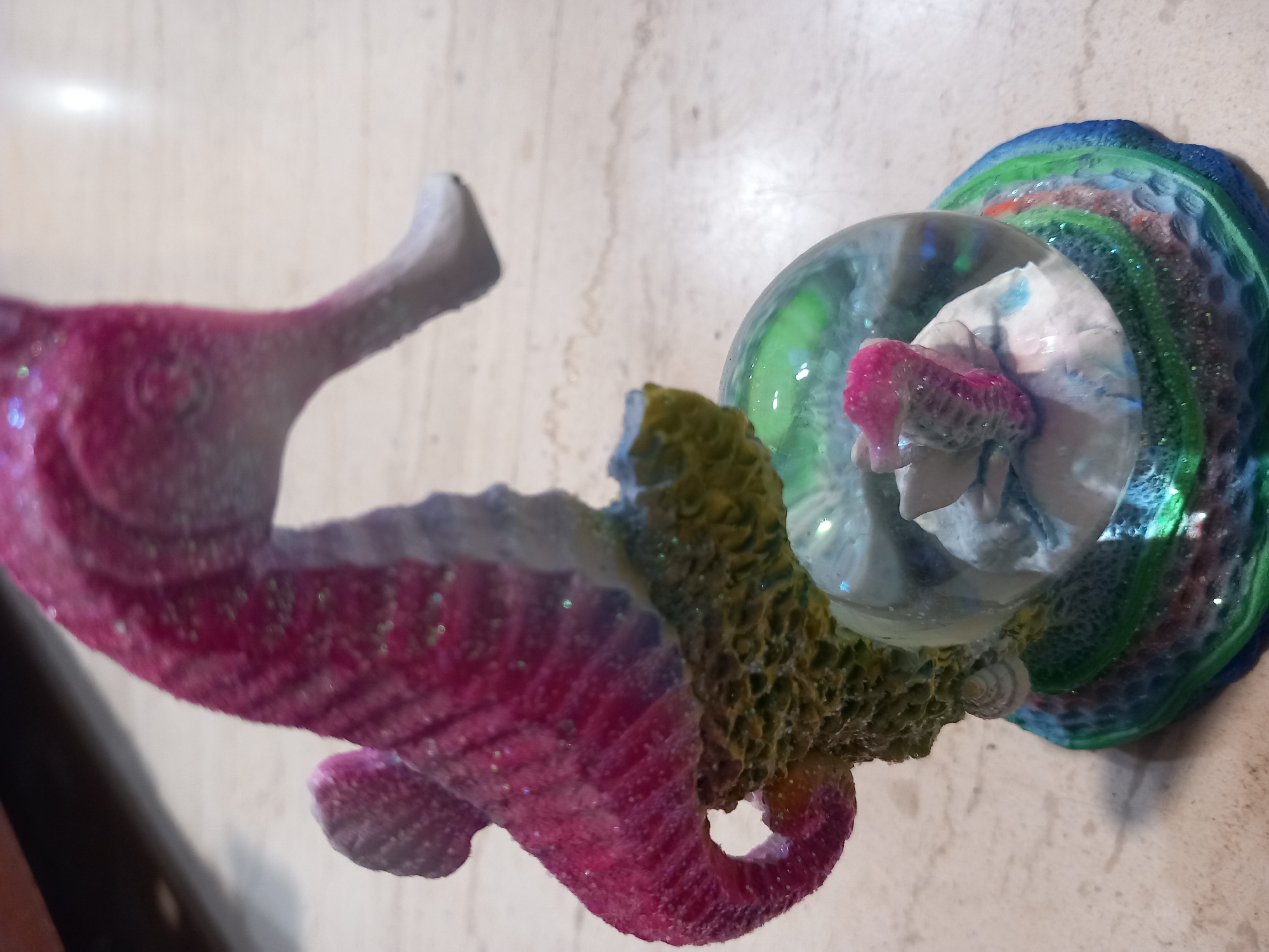 This media file features a close-up, angled shot of a decorative seahorse figurine. The seahorse is primarily pink and purple, with a shimmering glitter effect. It appears to be attached to a clear glass globe, which contains a small, pink, seahorse-like object inside, possibly another miniature figurine. The globe is supported by a textured, multi-colored base that resembles coral or a reef, with shades of blue, green, and yellow. The background is a light-colored, speckled surface, possibly a countertop or table. The lighting suggests it is daytime, and the focus is entirely on the figurine. There is no text visible in the image. The overall impression is of a whimsical, possibly underwater-themed, decorative item.