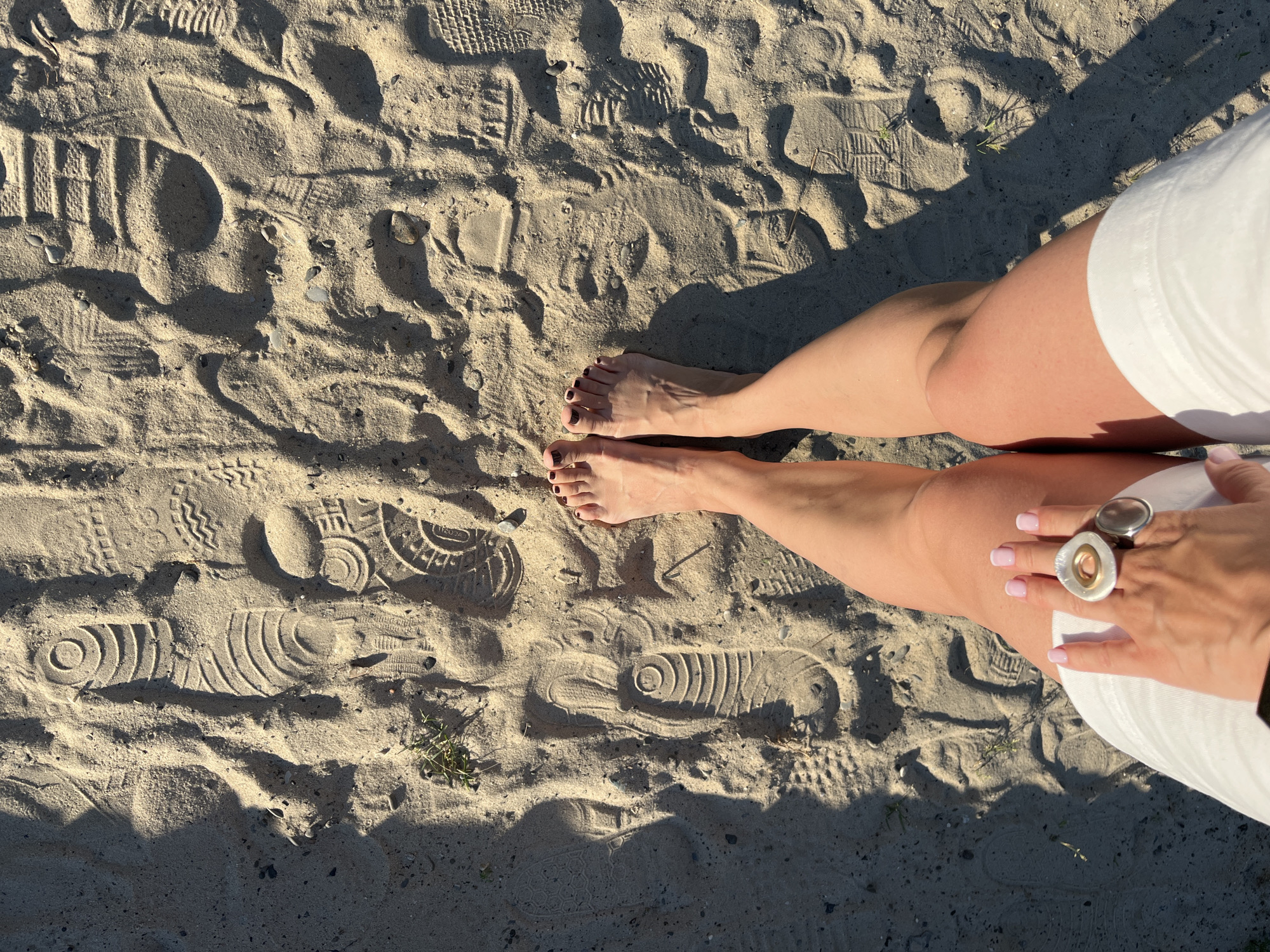 The image shows a pair of bare feet standing on sandy ground with many shoe imprints. The person is wearing white shorts and has painted toenails. Their right hand is visible, adorned with two rings, and manicured fingernails. The sunlight casts distinct shadows on the sand, suggesting a sunny day. The presence of shoe prints indicates this is likely a public area where people walk, such as a beach or park. The scene is likely taking place in Sochi, Russia, given the provided location context. The overall impression is one of relaxation and enjoying a warm, outdoor environment.