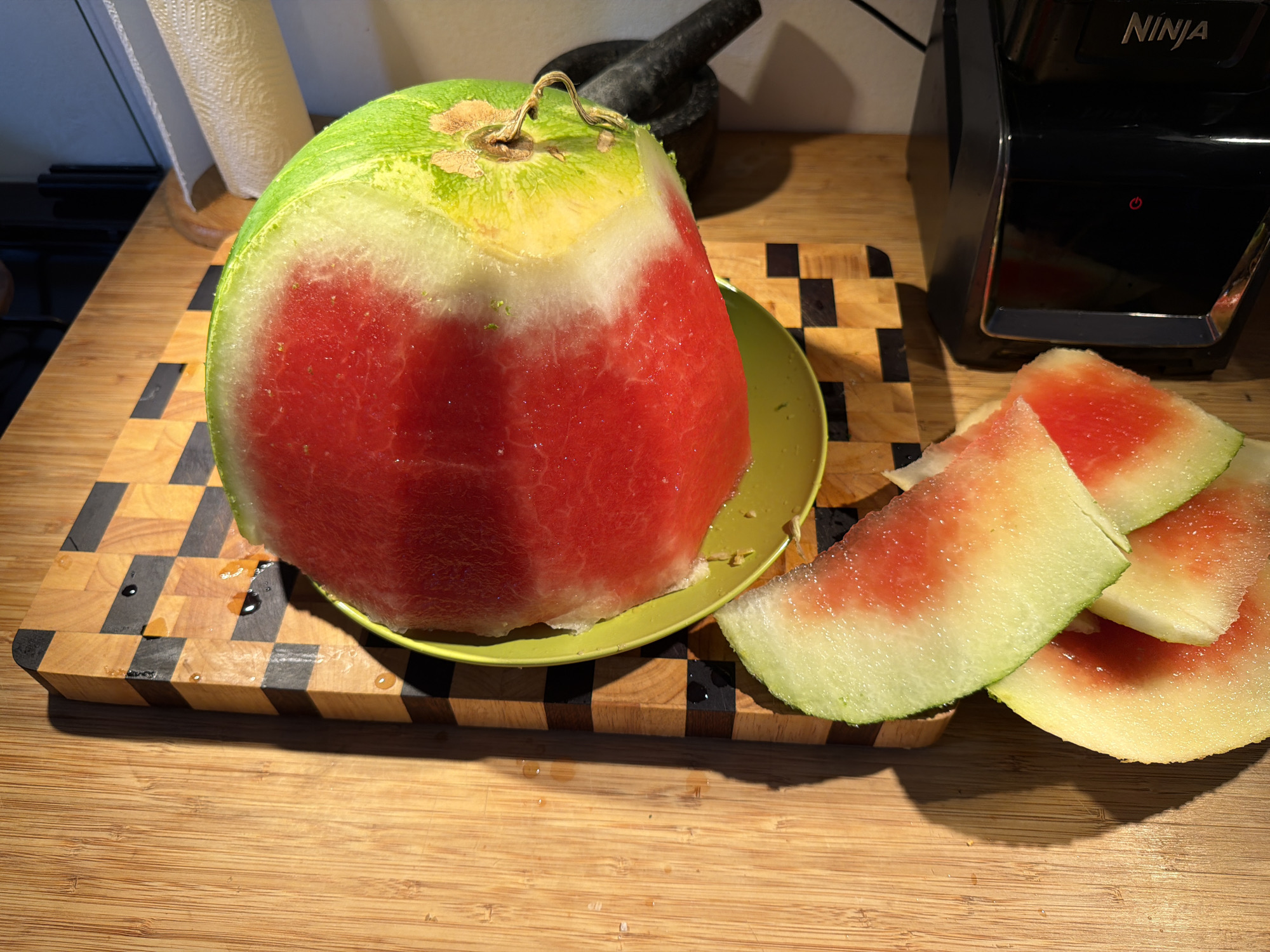 A close-up, high-angle shot shows watermelon rinds and pulp on a wooden cutting board. The watermelon pieces are arranged in the lower half of the frame, with the largest piece resting in a bright green bowl. The cutting board features a checkered pattern of light and dark wood. To the right of the cutting board, a black appliance, possibly a blender or juicer, is visible. In the background, a roll of paper towels and a dark object, possibly a mortar and pestle, are out of focus. The lighting suggests a natural light source, creating shadows on the wooden surface. The scene is set indoors, in what appears to be a kitchen environment. The location in Even Yehuda, Israel, is not explicitly depicted in the image itself.