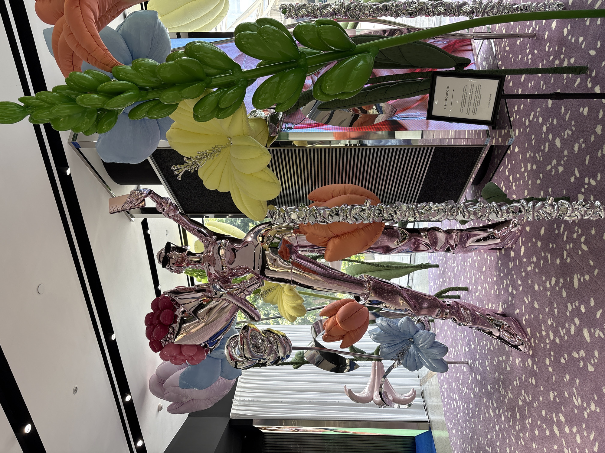 The image depicts an indoor art installation, seemingly located in Bangkok, Thailand, as indicated by the "GENTLE MONSTER" sign. The installation features a chrome-colored, life-size human figure surrounded by large, plush, pastel-colored flowers. These flowers are mostly variations of blues, yellows, oranges, and pinks, and some have metallic, twisted stems. The figure appears to be stepping forward or flying through the air, surrounded by the artificial floral display. Behind the figure is a display cabinet with a horizontally striped front and a screen displaying blurred imagery. Adjacent to this display is a sign that reads "AI PHOTO BOOTH." The floor is purple with white speckles. The wall to the left has black and white stripes.