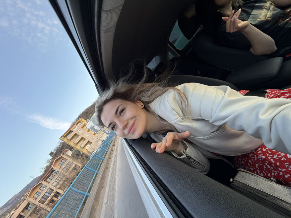 The scene depicts two individuals inside a moving vehicle, observed from an interior perspective looking out the front passenger window. The primary subject, a young adult female with blonde hair, occupies the front passenger seat. She wears a light-colored (likely white or cream) quilted leather jacket and a red and white floral-patterned dress or skirt. Her hair is windblown, and she smiles, looking towards the upper left. Her right hand is raised, fingers slightly splayed, near the open window. A light-colored purse with a patterned design rests on the seat beside her. In the rear passenger seat, a second individual is partially visible, wearing a dark t-shirt with a light graphic. This individual holds what appears to be a lit cigarette or similar object between the fingers of their raised right hand.
Outside the vehicle, a paved road or bridge is visible immediately next to a blue metal safety railing. Beyond the railing, several multi-story buildings of varying architectural styles are situated on a hillside, characteristic of the city of Divanovići, Montenegro. The hillside features sparse vegetation and some visible power lines. The sky is clear blue with wispy white clouds, indicating daylight conditions. The vehicle's interior shows dark upholstery and a visible sunroof mechanism at the top of the frame.