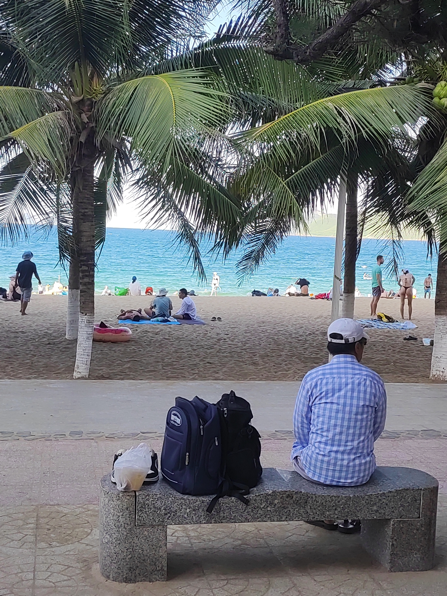 The image depicts a scene at a beach in Nha Trang Ward, Vietnam, likely during daylight hours given the bright lighting. In the foreground, a man wearing a blue and white checkered long-sleeved shirt and a white baseball cap is seated on a stone bench, facing away from the camera towards the beach and ocean. Two backpacks, one dark blue and the other black, are placed on the bench next to him, along with a plastic bag containing what appears to be a drink and a pair of shoes.
The beach itself is sandy, with several palm trees casting shadows. In the middle ground, various individuals are spread out on the sand, some lying down, others sitting. The ocean is visible behind them, a vibrant turquoise blue, with a distant shoreline or landmass visible on the horizon. A few people are standing near the water's edge. The overall atmosphere suggests a relaxed, possibly tourist-oriented environment.