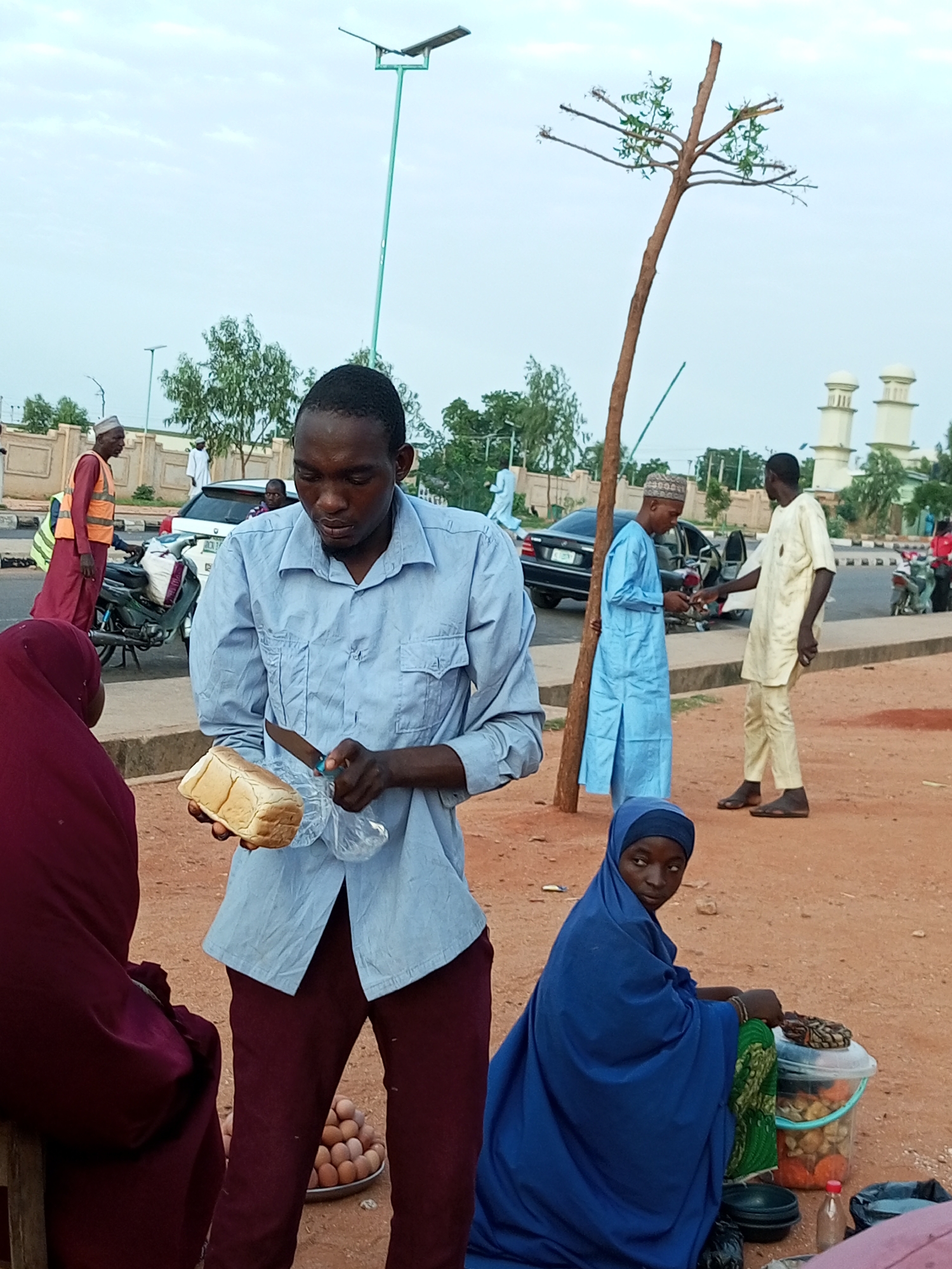 The image depicts an outdoor market scene in Gombe, Nigeria. In the foreground, a man wearing a light blue shirt and maroon trousers is slicing a loaf of bread with a knife. He is interacting with a person partially visible on the left, who is wearing a dark red hijab. Behind the bread seller, a pile of eggs is visible on a metal plate. To the right of the bread seller, a young woman in a blue hijab and green garment is seated, tending to goods in a clear plastic container.
In the mid-ground, several other individuals are present. A man in a light blue robe and a patterned cap is standing near a tree, seemingly in conversation with another man in a beige tunic and trousers. Further back, a man in an orange vest and maroon trousers is standing near a motorcycle. Several vehicles, including a white SUV and a black car, are parked on the side of the road.
The environment is an unpaved area with scattered vegetation and a cloudy sky. A street light on a tall pole is visible in the background, along with the distinctive domes of a mosque. The scene appears to be set along a roadside, suggesting a busy commercial area within the city of Gombe.