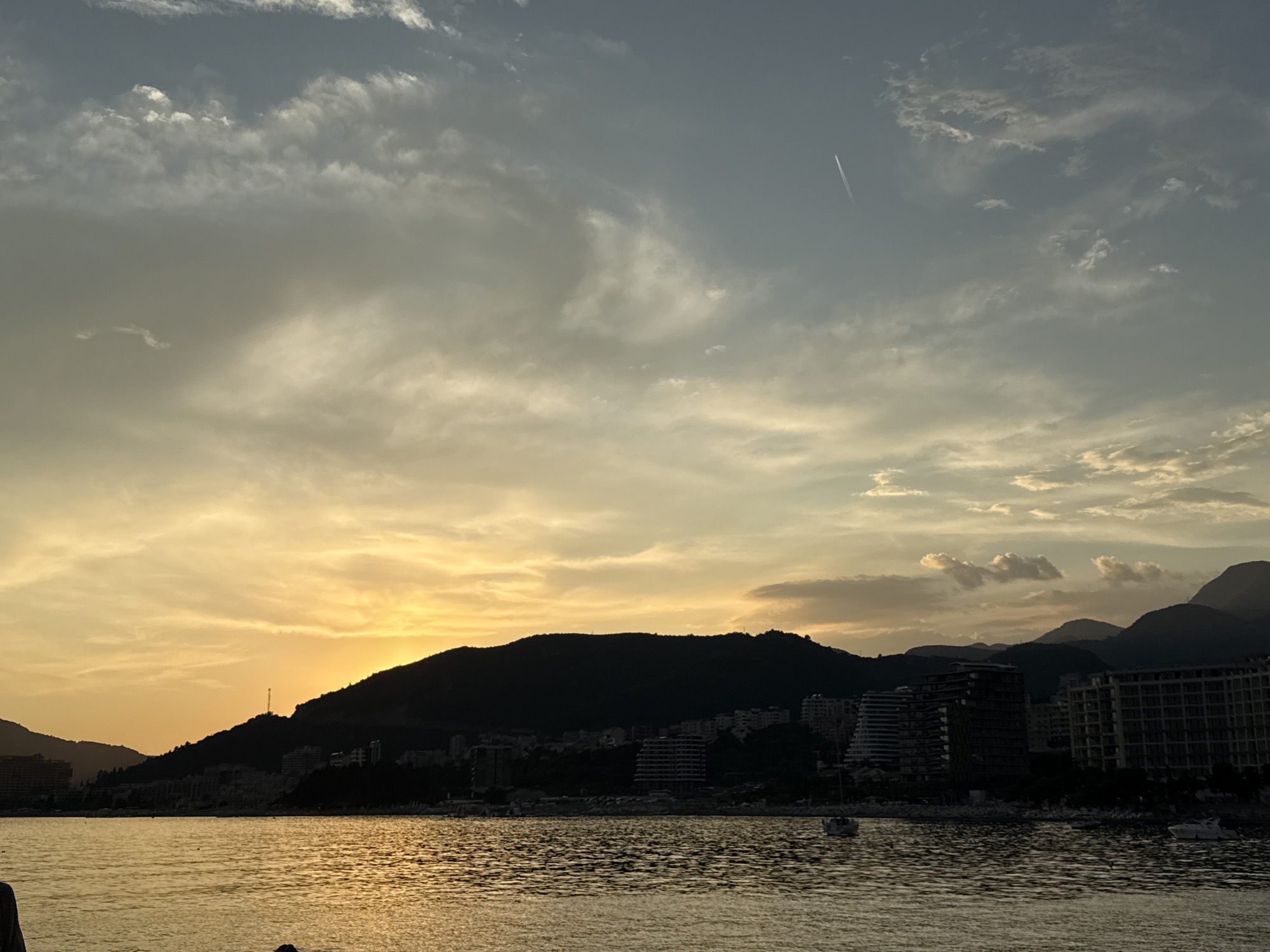 This media file captures a serene sunset scene over Bečići, Montenegro. The foreground is dominated by a calm body of water, reflecting the warm hues of the setting sun. The water's surface is gently rippled, creating a shimmering effect.
In the mid-ground, a silhouetted hillside is visible, dotted with buildings that form a coastal town. These structures are dark against the bright sky, suggesting they are either set back from the shore or facing away from the light. Several modern buildings, possibly hotels or apartment complexes, are identifiable with their distinct architectural designs. A prominent tower or antenna can be seen on top of the hill.
The background features a majestic mountain range, also in silhouette, with multiple peaks fading into the distance. Above the land, the sky is a canvas of dramatic clouds. The lower part of the sky, near the horizon, is awash with vibrant yellow and orange tones, indicative of sunset. Higher up, the clouds transition to softer grays and whites, with a few wispy formations adding texture. A faint contrail from an airplane streaks across the upper right portion of the sky, adding a touch of human presence to the natural spectacle.
The overall mood is peaceful and tranquil, evoking a sense of calm and the beauty of nature at dusk. The time of day is clearly sunset. The weather appears clear with scattered clouds. The presence of the sea, coastal town, and mountains strongly suggests a seaside resort location, consistent with Bečići, Montenegro. There is no visible text in the image.