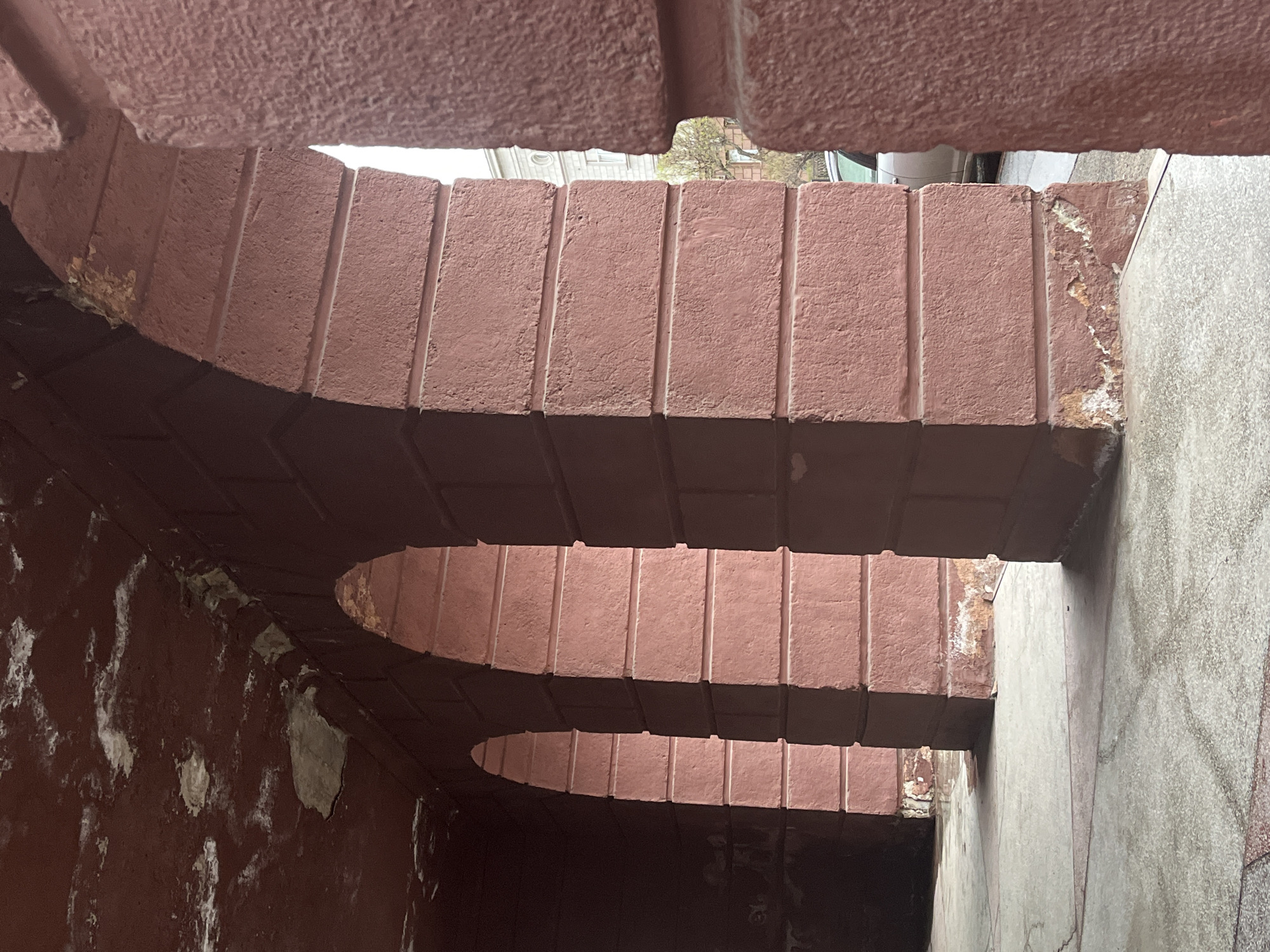 An interior shot shows the underside of a weathered, tiered archway. The arches are constructed from a pinkish-red, textured material, possibly brick or concrete, and feature vertical grooves along their surfaces. The walls on either side of the archway exhibit signs of age and deterioration, with peeling paint and discoloration. The overall lighting is dim, highlighting the contrast between the textured surfaces and the shadows within the archway. The exterior is visible through the arches.