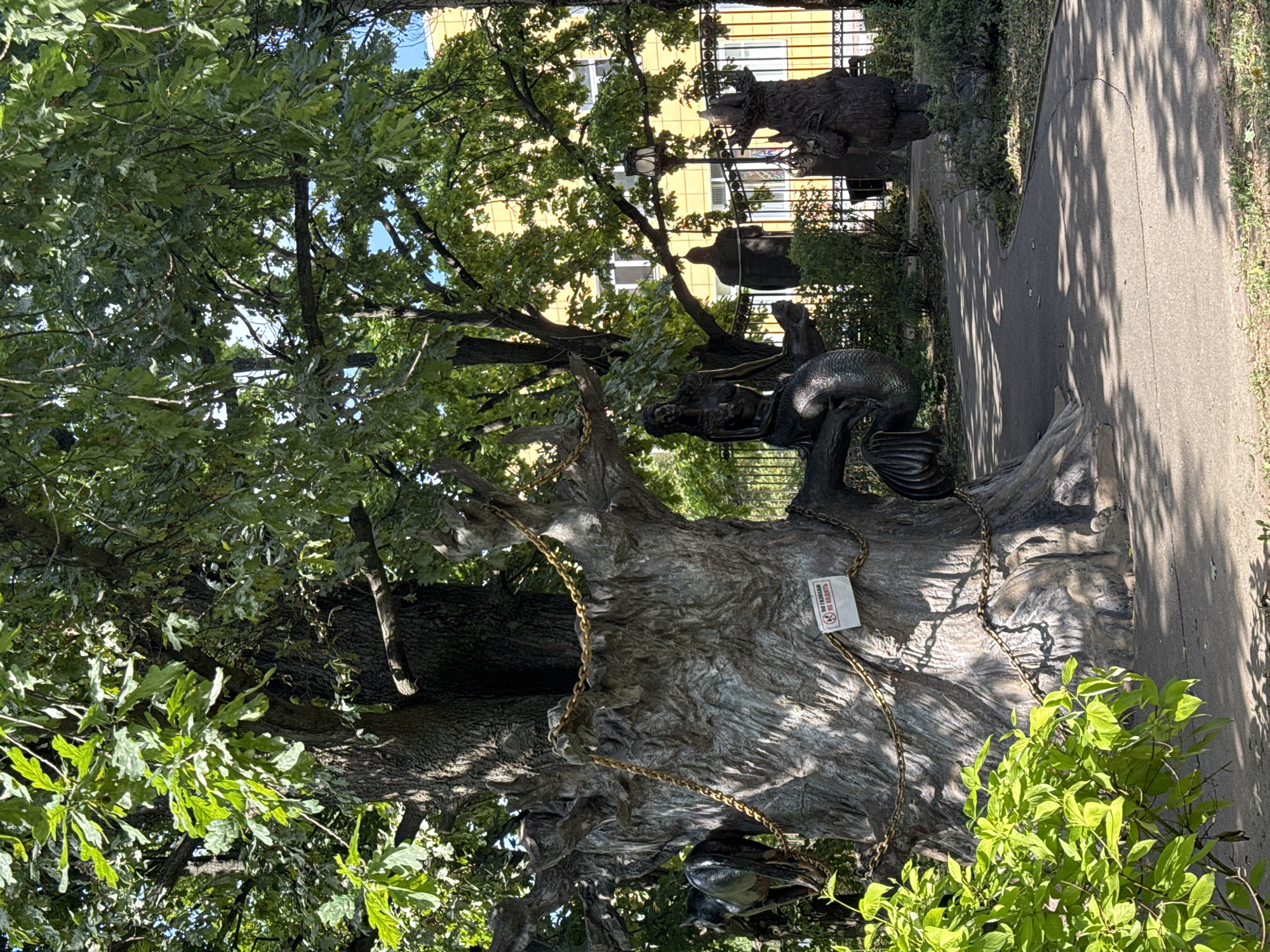This image shows a large, ancient oak tree in the city of Sovkhoza imeni Lenina, Russia. The tree is adorned with large, thick gold chains wrapped around its trunk and branches. A sign reading "NO SMOKING" is affixed to the trunk. To the left of the tree, a statue of a black horse or similar animal is partially visible. On a branch of the tree, there is a bronze sculpture of a mermaid. Further back, a pathway winds through the park, flanked by manicured bushes and trees. In the background, a yellow building with windows is visible behind a black metal fence. Other statues, including one of a human figure and another resembling a bear, are scattered throughout the park. The ground is paved with concrete, and dappled sunlight filters through the leaves of the trees, creating shadows on the path.