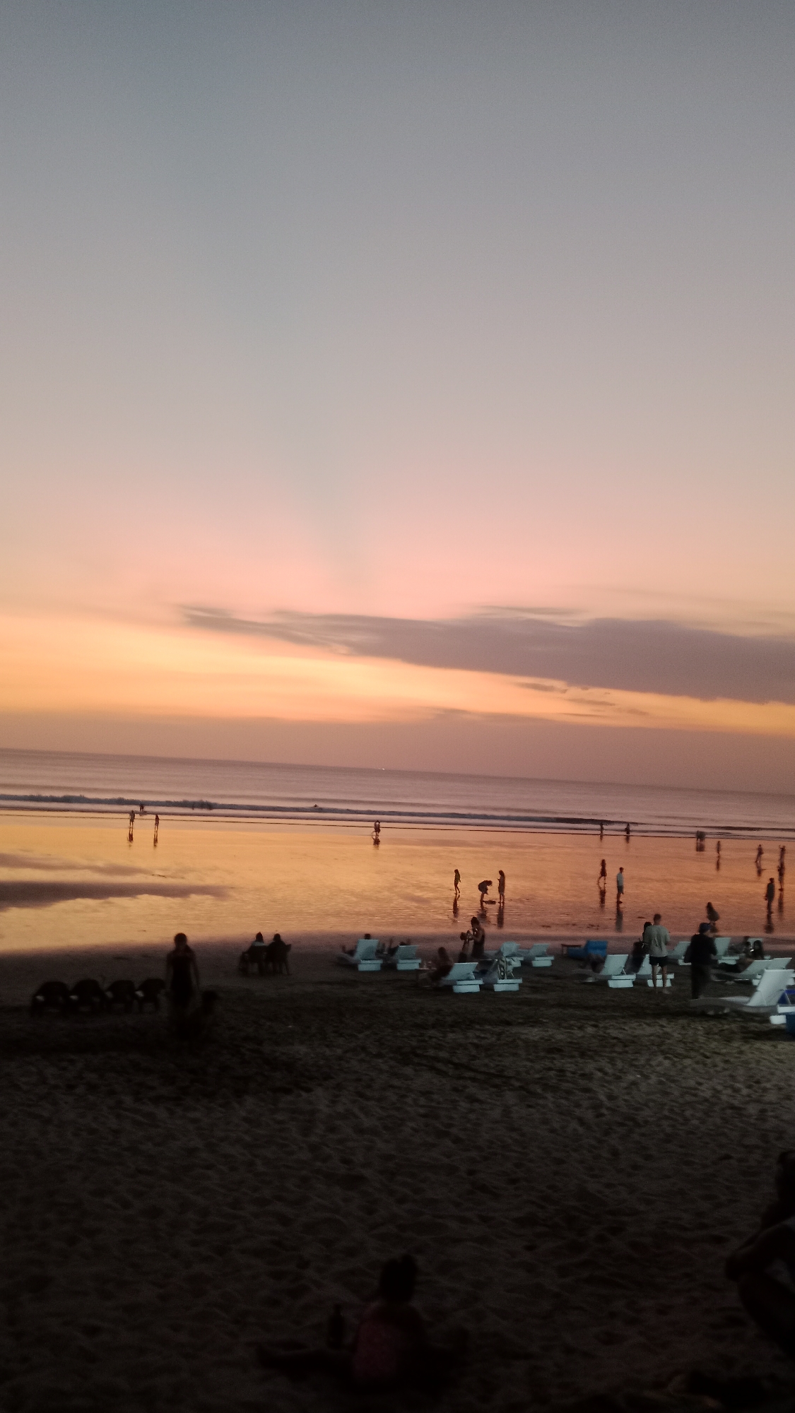 The image depicts a serene sunset scene on Kuta Beach in Bali, Indonesia. The sky transitions from a pale gray at the top to warm hues of orange and pink near the horizon. The ocean, reflecting the colors of the sky, is calm with gentle waves lapping at the shore. The wet sand of the beach is also reflective, creating a mirror-like effect.
Several people are visible scattered across the beach and in the shallow water. Some are walking, while others appear to be interacting or relaxing. In the foreground, closer to the viewer, are dark silhouettes of people sitting on the sand, some appearing to be children. Further back, on the beach, are lounge chairs, some occupied by individuals, suggesting a relaxed atmosphere. Groups of people are walking near the water's edge, and a few are wading in the sea. The overall impression is one of a peaceful evening gathering at a popular tourist destination. The lighting is dim, characteristic of dusk, with the warm glow of the sunset dominating the scene.