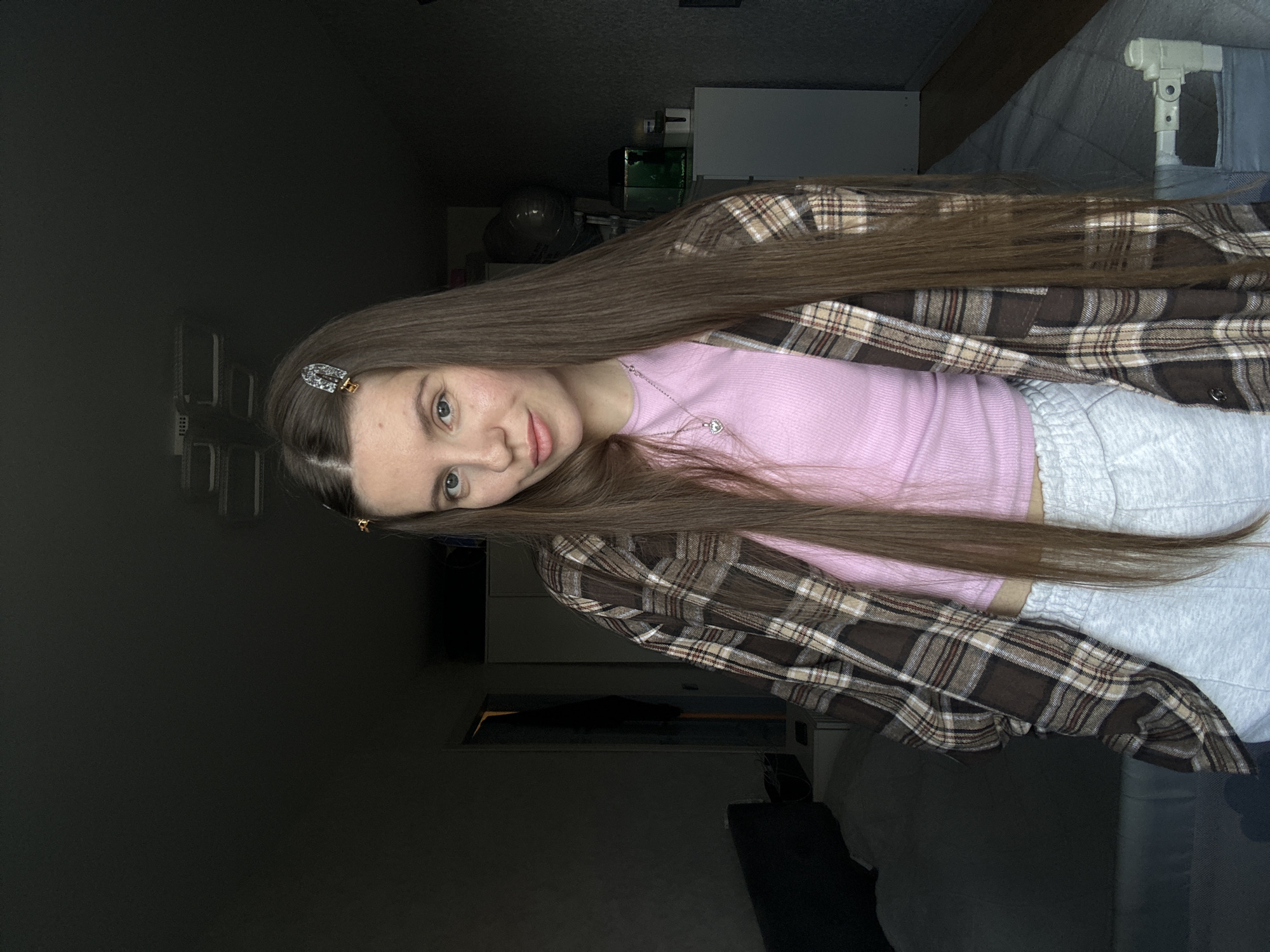 A woman with long brown hair poses indoors. She wears a pink top under a brown and beige plaid flannel shirt, along with light gray sweatpants. Her hair is held back with hair clips. The background shows various objects including a humidifier, possibly a closet, and a bed. The lighting is somewhat dim. The location in Klin, Russia is not determinable from this image.