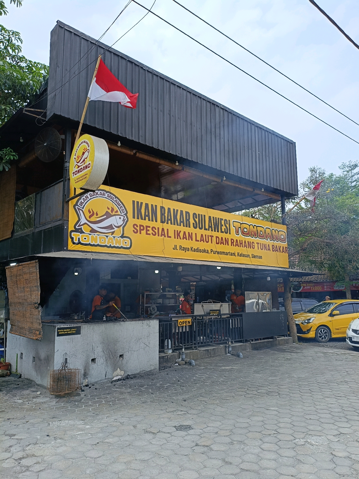 The image shows a restaurant called "Tondano" in Maguwoharjo, Indonesia, specializing in grilled fish from Sulawesi. The restaurant has a prominent yellow banner with the text "IKAN BAKAR SULAWESI TONDANO" and "SPESIAL IKAN LAUT DAN RAHANG TUNA BAKAR." Below the banner, the address "Jl. Raya Kadisoka, Purwomartani, Kalasan, Sleman" is visible.
The restaurant appears to be an open-air establishment with a grilling area where a person in an orange shirt is tending to food. There is smoke emanating from the grilling area. A red and white Indonesian flag is displayed on a pole above the restaurant. In front of the restaurant, there is a cobblestone paved area with several vehicles parked, including a yellow car. The environment suggests a warm, possibly humid, climate with lush greenery visible in the background.