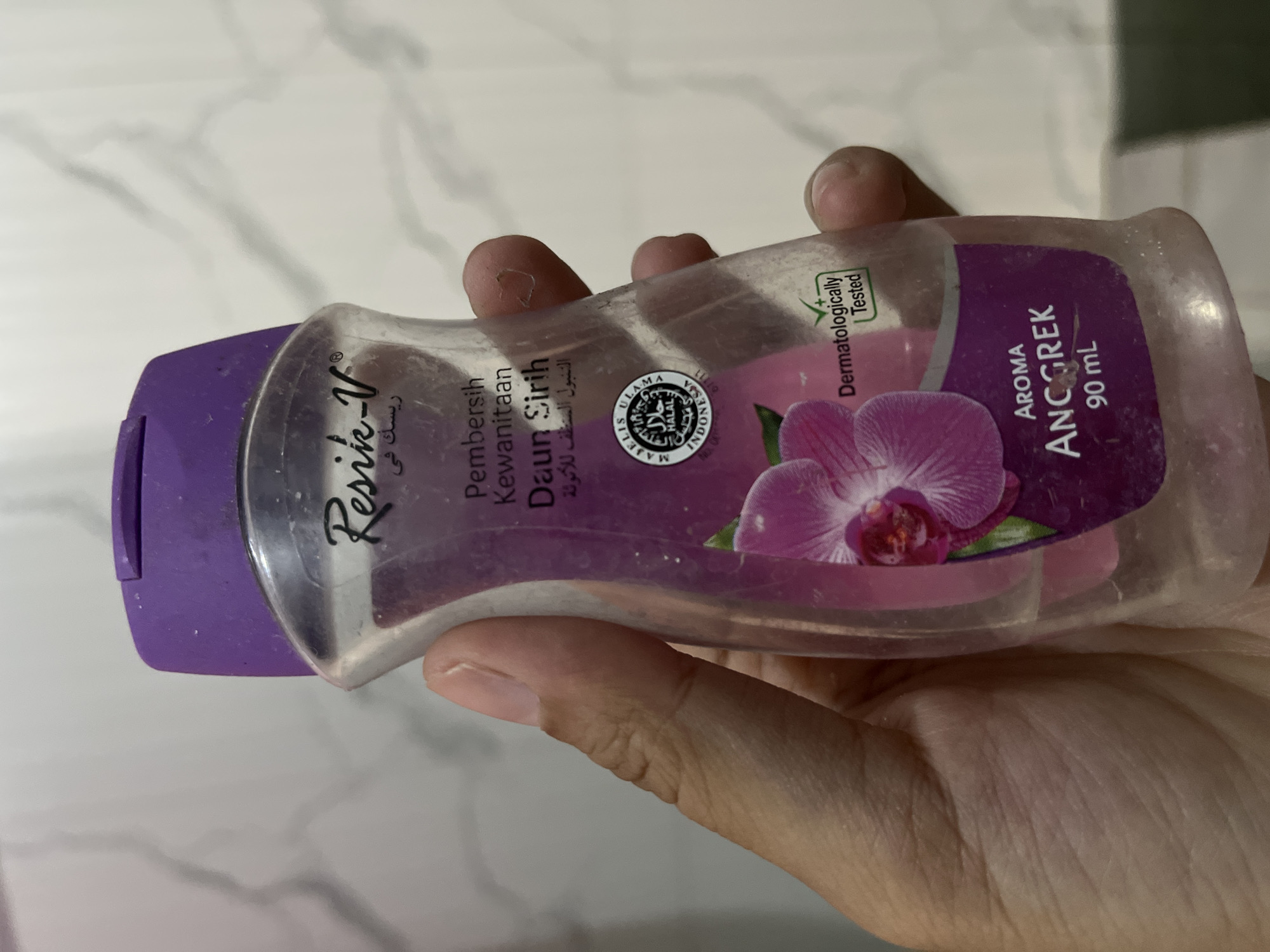 A person's hand holds a transparent plastic bottle with a purple cap and a purple label. The bottle contains a pink liquid and has a label with text and an image of a pink orchid. The text on the label includes "Resik-V", "Pembersih Kewanitaan", "Daun Sirih", and "Aroma Anggrek 90 ml". There is also a Halal certification logo and a "Dermatologically Tested" logo. The background appears to be a marble-patterned wall. The image is taken in Kalisampurno, Indonesia.