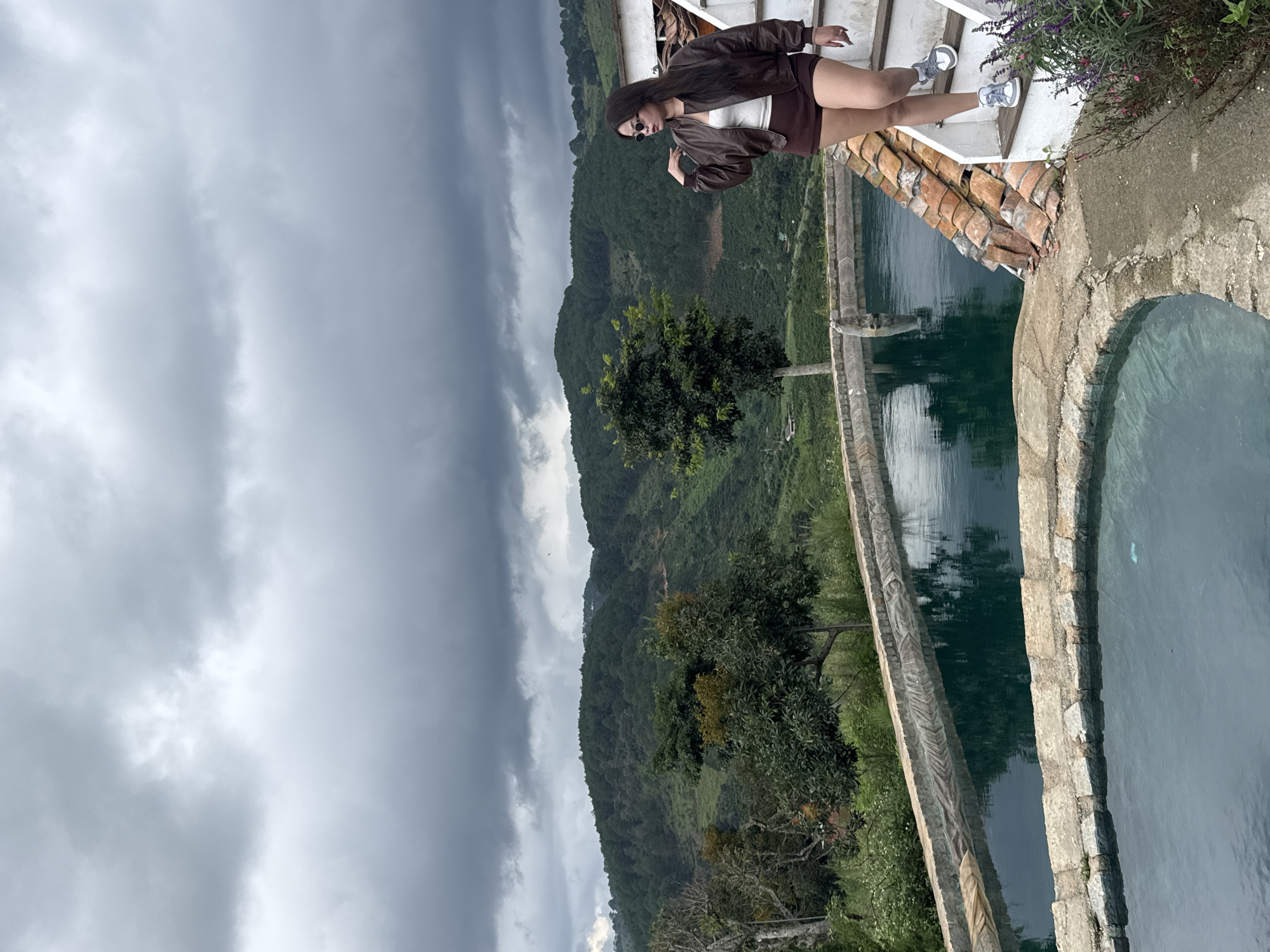 A young woman with long dark hair stands on a white staircase with terracotta tile accents, looking to her left. She is wearing a brown leather jacket, a white top, and brown shorts. She has on sunglasses and white sneakers. Behind her, a scenic mountain landscape is visible under a cloudy sky. In the foreground, there are two large, stone-lined pools of dark green water. The woman appears to be at an outdoor location, possibly a resort or private residence, in Thôn 3, Vietnam, given the mountainous terrain and vegetation.