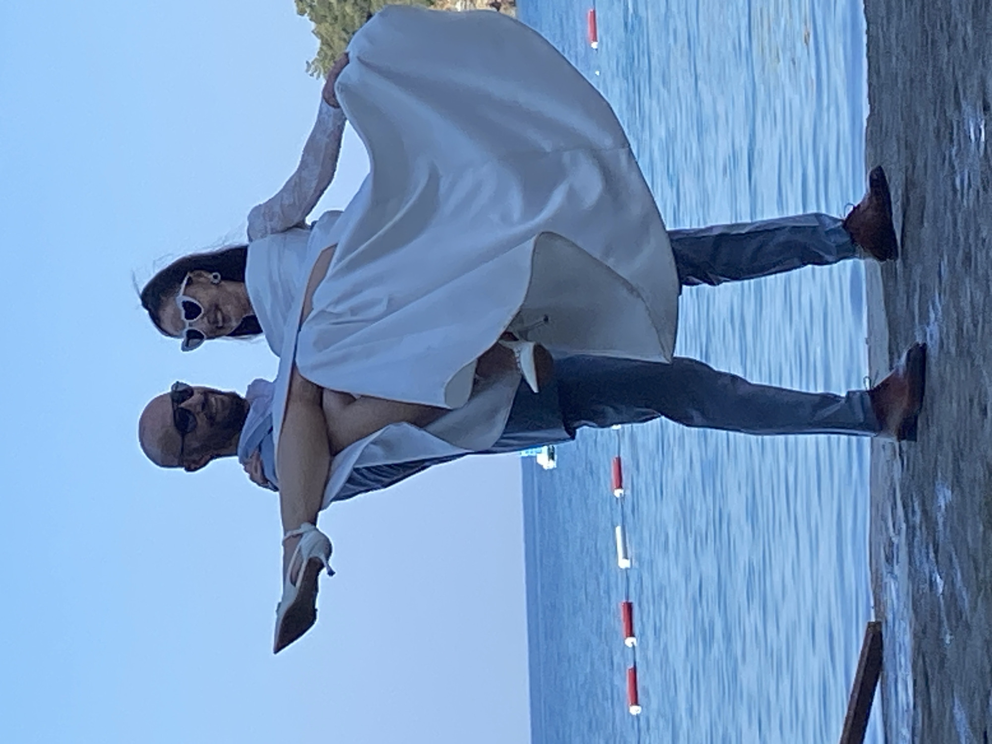 In the city of Budva, Montenegro, a man carrying a woman on a pier by the sea stands with the woman's white wedding dress flowing around them. The couple is smiling and wearing sunglasses. The man has a bald head and is wearing a blue suit and brown shoes. The woman is wearing a white wedding dress with lace sleeves and white high-heeled shoes. The sea is blue and calm, with buoys floating in the water. Trees can be seen on the shore in the background. The sky is clear and blue.