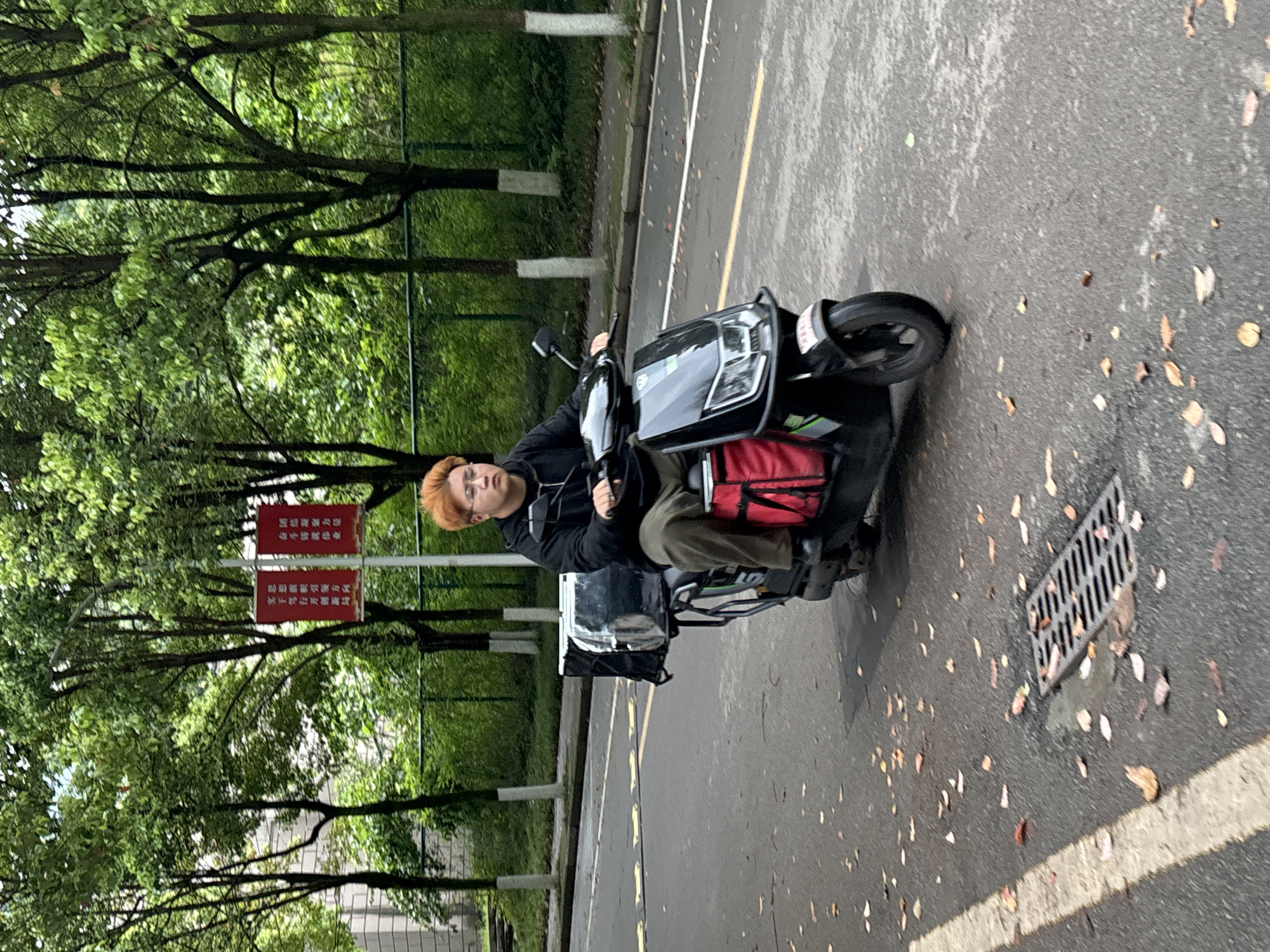 The image shows a young man sitting on a black electric scooter on an asphalt road. He is wearing a black hoodie and green pants. He has orange hair and is looking directly at the camera. The scooter has a red bag attached to the back and a white box.
The environment appears to be a park or a street with trees lining the side. The trees have white paint around the trunks. There are some road markings and a metal drain cover visible on the road. There are red signs with white Chinese writing on them. Based on the text on the red signs, the location is likely in the city of Jiangjin, China.