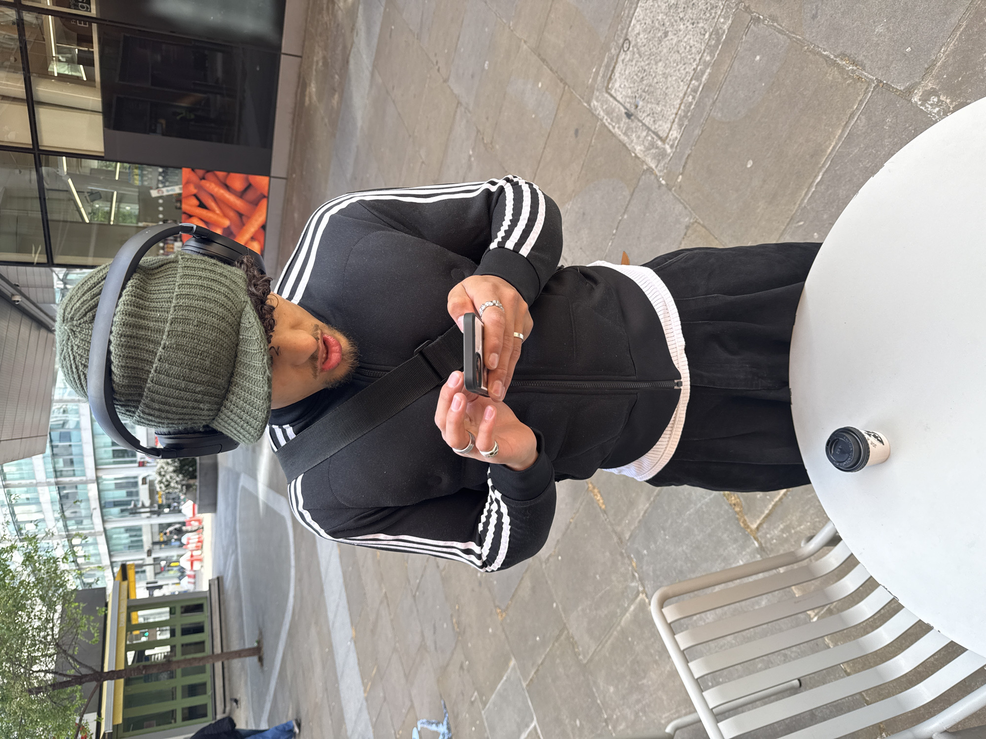 The image depicts a young man in an outdoor urban setting, likely a pedestrian area in London. He is standing or leaning near a white round outdoor table with a light grey slatted metal chair next to it.
The man is wearing a black Adidas tracksuit featuring prominent white stripes along the sleeves and sides of the trousers. He has a green knit beanie on his head and black over-ear headphones are worn on top of the beanie. He possesses dark curly hair and a beard or stubble. He is holding a smartphone in both hands, looking down and seemingly engrossed in its screen. Several rings adorn his fingers, including two on his left hand and a plain gold band on his right. A black shoulder bag strap is visible across his body.
On the white table, there is a small, white disposable cup with a black lid. The text "30ML" is clearly visible on the side of the cup, with some additional, partially obscured text on the black lid.
The background showcases a modern city environment with tall, glass-fronted buildings, one of which reflects the surroundings. A digital display or billboard behind the man prominently features a vibrant image of carrots. To the left, a small tree with green foliage grows from the paved ground. Further in the background, there is a structure resembling a green kiosk or small building. The ground is paved with rectangular stone or concrete slabs in varying shades of grey. The overall lighting indicates daytime with bright, even illumination.