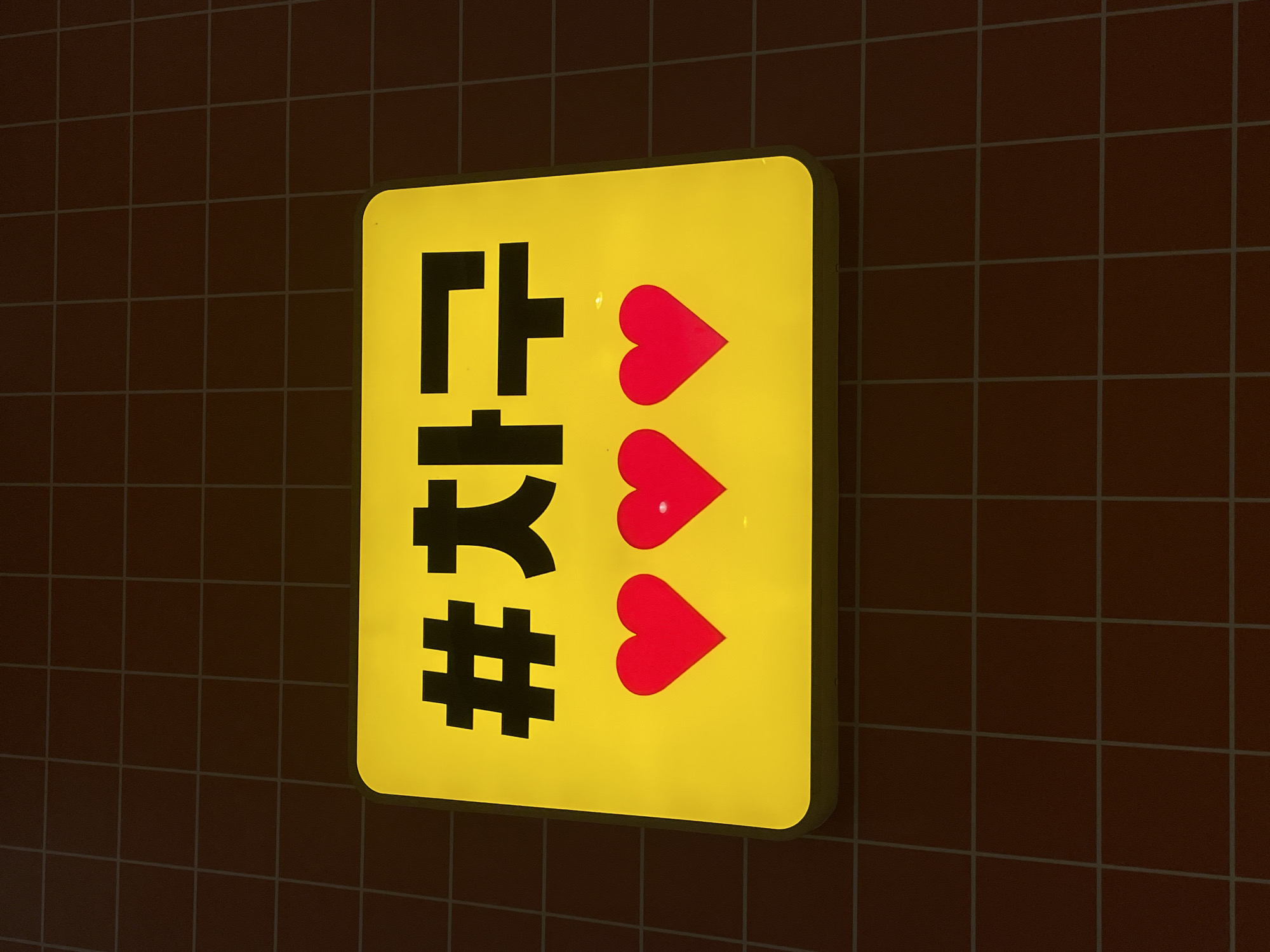 A close-up, eye-level shot showcases a bright yellow, illuminated rectangular sign against a dark, grid-patterned background. The sign features black Korean characters that read "#차구" (which translates to "#Chagu" or "#Chagu-gu"). Below the text are three vibrant red heart shapes, equally spaced. The sign is the primary focus, with the textured background providing a contrasting backdrop. The overall lighting suggests it might be nighttime or an indoor setting with controlled lighting. There are no people or animals visible in the image, nor is there any indication of the specific location in Denpasar, Indonesia.