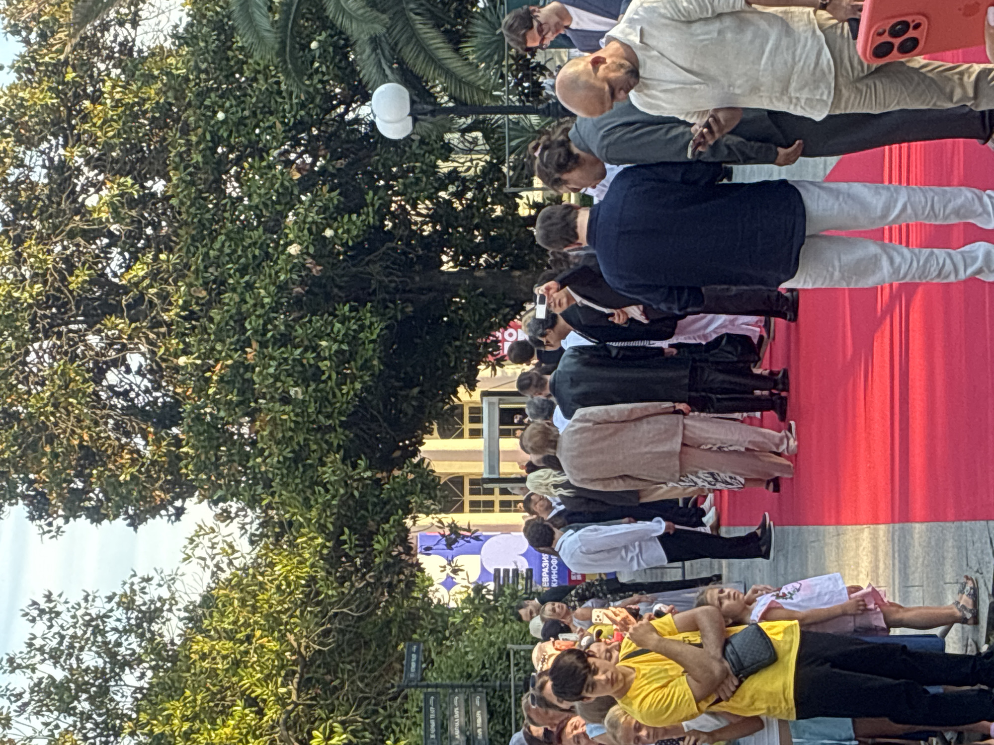 An outdoor scene in Sochi, Russia, captured on a red carpet event. A crowd of people, dressed in formal and semi-formal attire, are gathered. The focal point is the red carpet, which extends into the distance. Trees and palm trees form a lush green backdrop, with a building featuring large windows visible in the background. There are signs in Russian language along the left side of the image. Several individuals are holding up their phones, suggesting they are taking pictures or recording the event. A young boy in a yellow t-shirt stands prominently on the left, and a young girl in a pink dress is beside him. The lighting suggests it is late afternoon or early evening.
