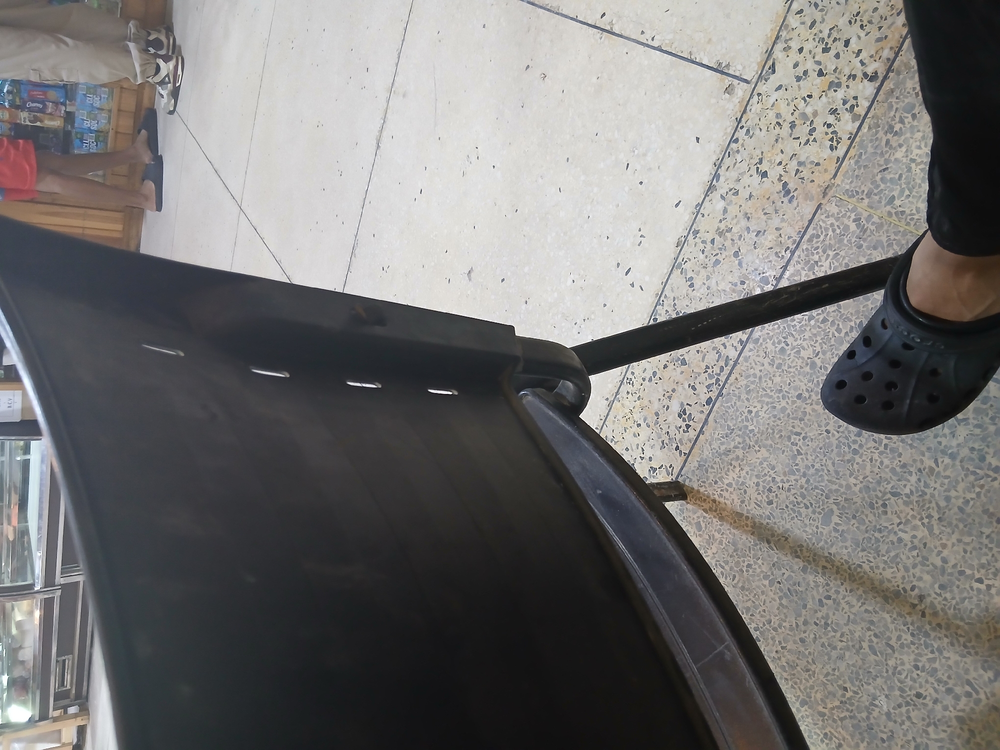 A person's left foot wearing a black clog is visible in the foreground, resting on a speckled floor made of tiles. Behind the foot, a dark plastic chair is partially visible, with its back and legs dominating the mid-ground of the image. In the background, a display counter with glass cases is set up, suggesting a commercial establishment. Two other individuals are partially visible in the background, one wearing red shorts and black sandals, and the other wearing light-colored pants and sneakers. The scene appears to be an indoor setting, possibly a store or a waiting area, with a tiled floor and display units common in commercial spaces. The lighting suggests it is daytime. The location of Santa Ana, Venezuela, is not directly evident from the image itself but is provided as context.