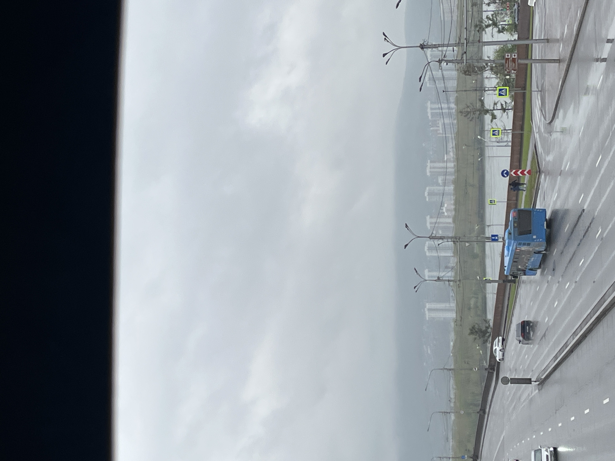 The image shows a street view in Krasnoyarsk, Russia on an overcast day. A blue bus is moving on the road. Several cars are also visible. There are street lights and pedestrian crossing signs alongside the road. High-rise buildings are visible in the background through the overcast sky. The road appears to be wet, suggesting recent or ongoing rain. A dark object is visible on the left edge, possibly a window frame.