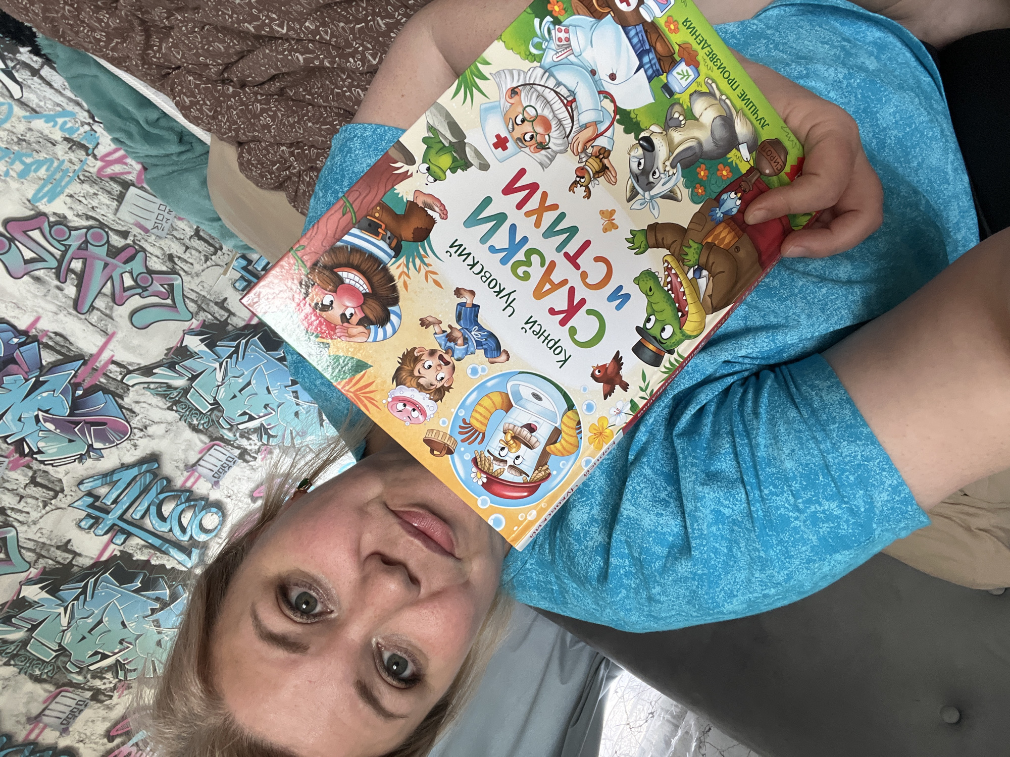 A woman, appearing to be middle-aged with light blonde hair and light-colored eyes, is captured in a high-angle shot, looking directly at the camera with a neutral expression. She is wearing a textured blue t-shirt and holding a colorful children's book. The book's cover features various cartoon characters, including a doctor, a wolf, a crocodile in a top hat, and a man with a large nose in a sailor suit. Prominently displayed Russian text on the cover reads "СКАЗКИ И СТИХИ" (Tales and Poems) by "Корней Чуковский" (Korney Chukovsky), with "ЛУЧШИЕ ПРОИЗВЕДЕНИЯ" (Best Works) also visible.
The woman is positioned on what seems to be a bed or sofa. The background to her left features patterned bedding with graffiti-style blue and pink lettering on a white brick-patterned surface. A dark gray upholstered furniture piece with two buttons is visible to her right. Other textiles, including a brown patterned fabric and a teal blanket or clothing, are also present in the background. The setting is indoors, characteristic of a casual, everyday moment. The image was taken in Vesyoloye, Russia.