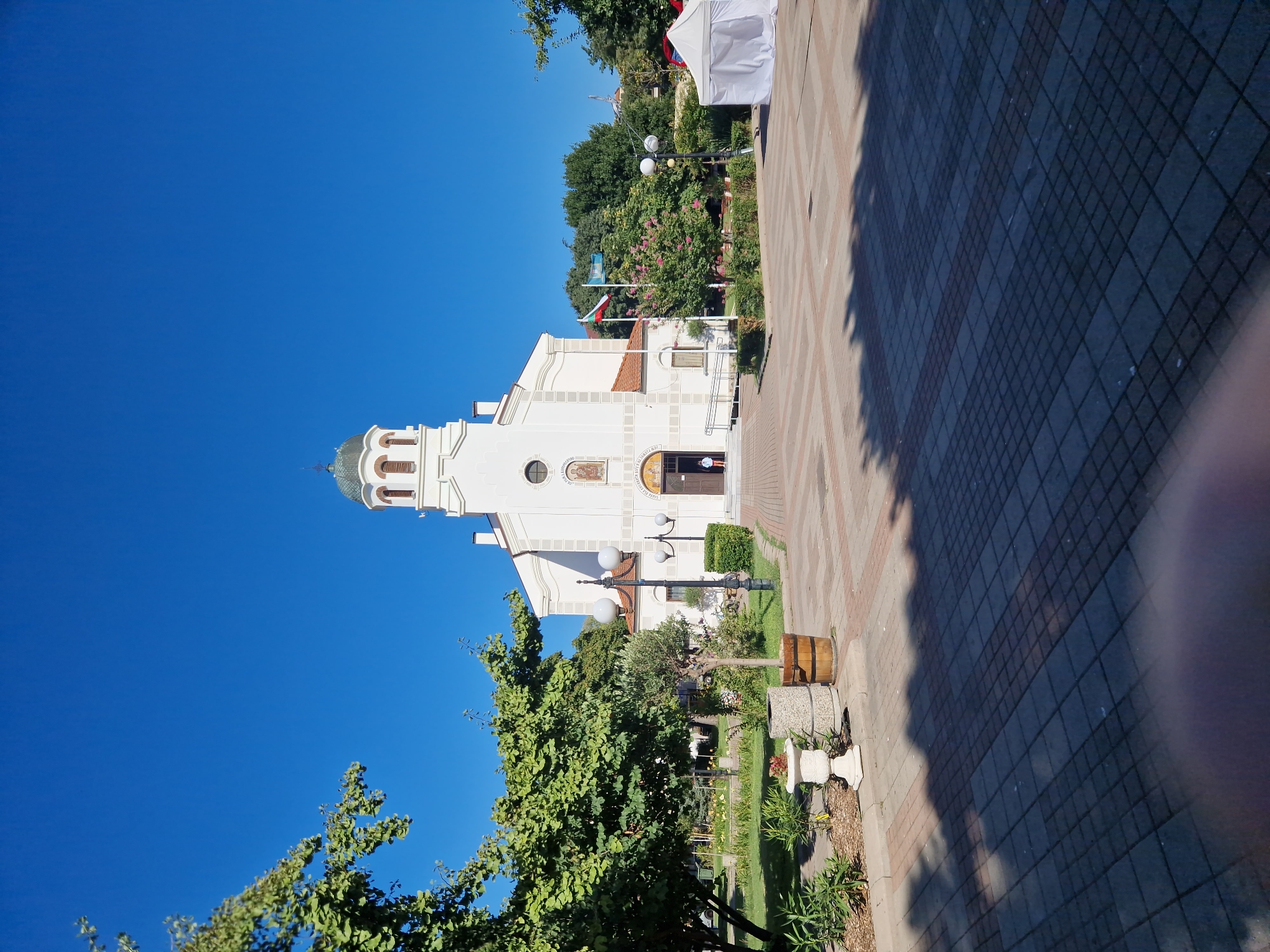 This is a medium-shot, eye-level, outdoor, vertical photograph of a white church in Pomorie, Bulgaria, on a clear, sunny day with a bright blue sky. The church has a dome-topped bell tower and a light-colored facade with decorative elements and religious artwork above the entrance. Two flags, one appearing to be the Bulgarian flag, are planted to the right of the church entrance. The church is situated in a paved courtyard with patterned brickwork, and there are trees and greenery surrounding the area. A white tent is visible in the background to the right. A single person, wearing a light-colored top and shorts, is standing near the open entrance of the church. The foreground shows a shadow cast by trees, creating a dappled effect on the pavement.