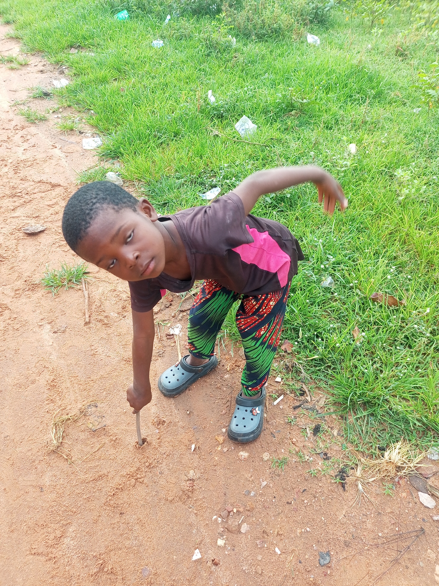 A young Black child is shown in a high-angle shot outdoors in Tempure, Nigeria. The child is wearing a brown t-shirt with a pink design and patterned leggings, as well as grey clogs with a ladybug charm. They are leaning forward, drawing in the reddish-brown dirt with a stick held in their right hand. The ground is a mix of dirt and sparse vegetation, with patches of green grass visible in the background, some of which is littered with white plastic. The child's left arm is extended upwards and to the right, and their gaze is directed towards the viewer.