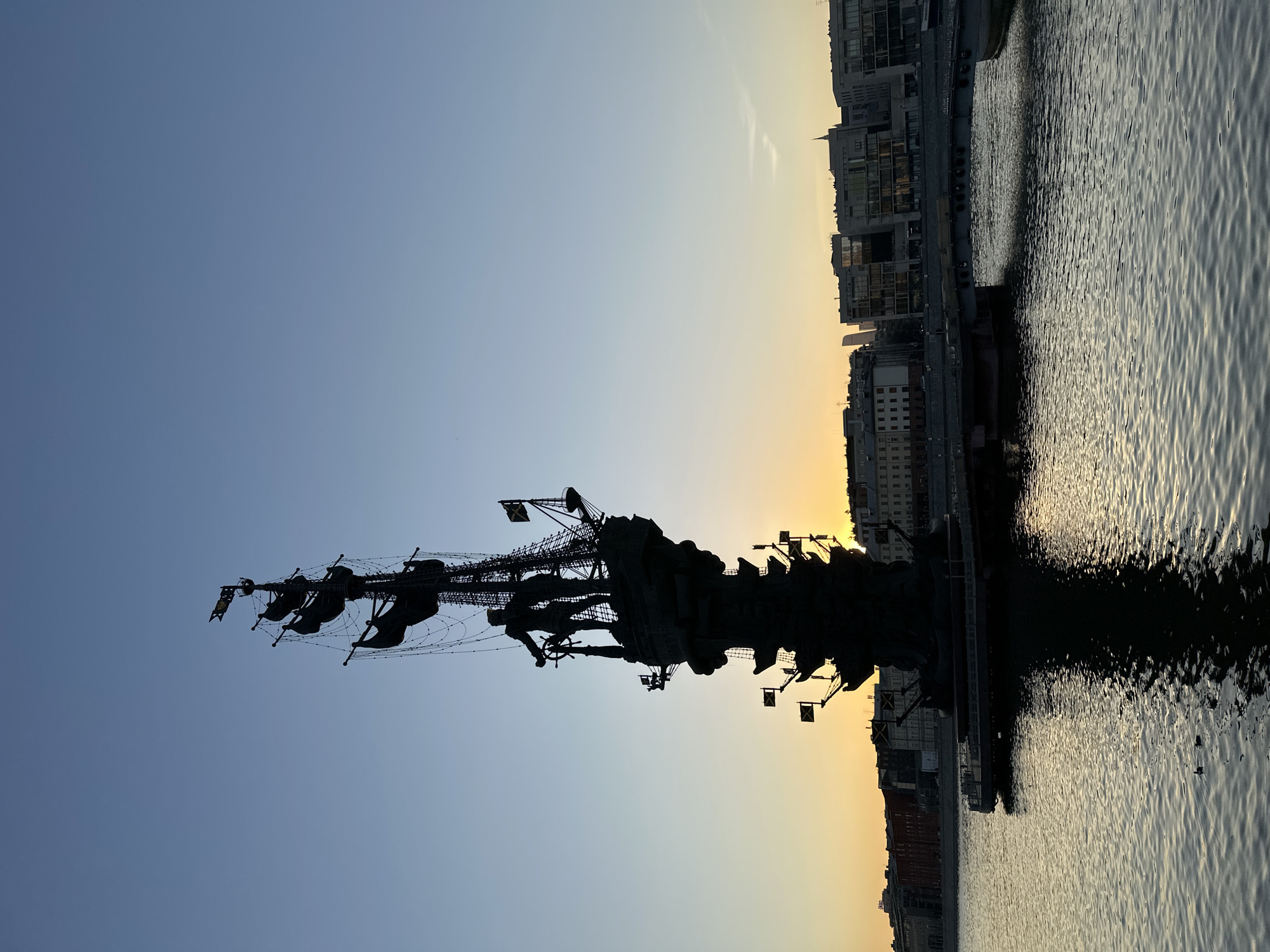 The image depicts a silhouette of the Peter the Great Statue in Moscow, Russia, set against a backdrop of a building-lined riverbank and a gradient sky transitioning from blue to orange at sunset. The statue, representing a ship with Peter the Great at its helm, is silhouetted. The river reflects the light, creating a shimmering effect.