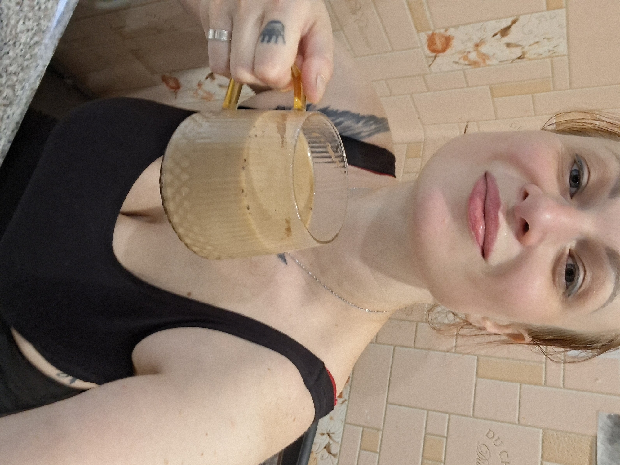 A woman is holding a glass mug of coffee. She is wearing a black tank top and a silver ring on her left hand. The background appears to be a tiled wall. The image is taken from the chest up, with the woman looking towards the camera.