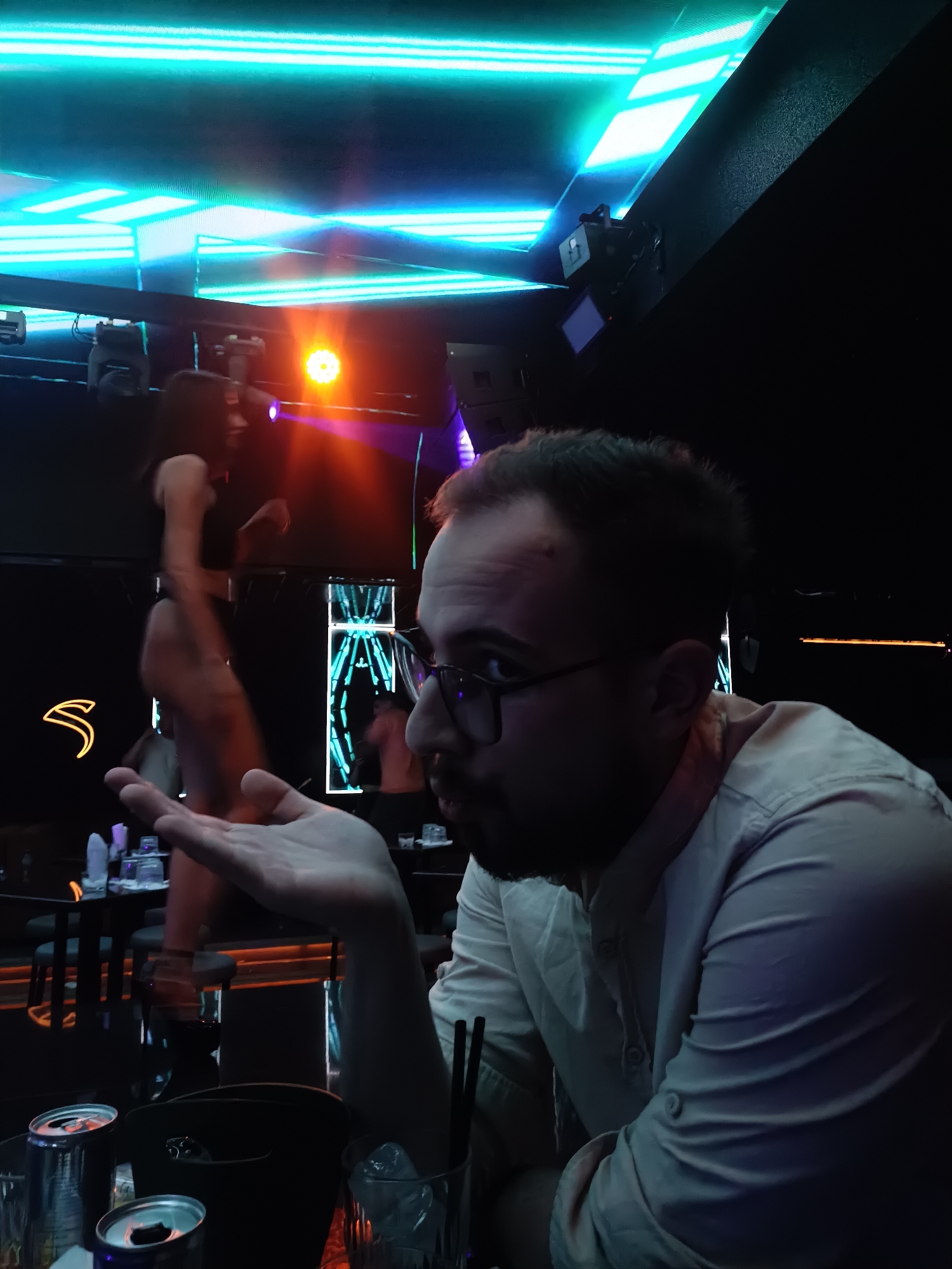 A man in a light-colored button-down shirt sits at a table with drinks, including cans, glasses with ice, and straws. He wears glasses and has a beard. His left hand is extended in a gesture. In the background, a dimly lit stage features a woman in a dark outfit and an illuminated backdrop. The location appears to be a nightclub with bright neon lighting in Plovdiv, Bulgaria.