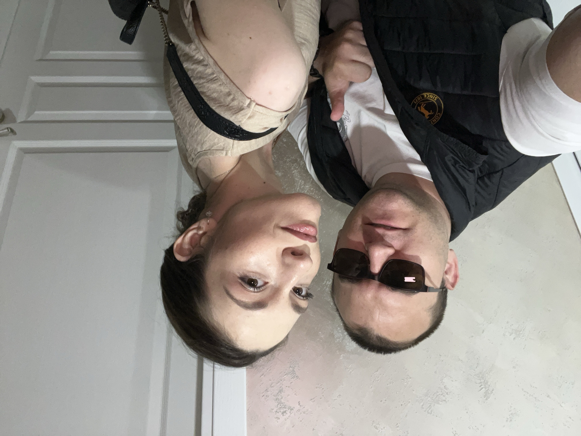 Two individuals are positioned closely together, facing the camera. The person on the left is a woman with dark hair and neutral makeup. To her left, a white door is visible. The person on the right appears to be a man wearing sunglasses, a white t-shirt, and a black vest with a gold emblem on the chest. The background behind them is an off-white wall with a textured or mottled appearance. The location is an Unknown city of Unknown.
