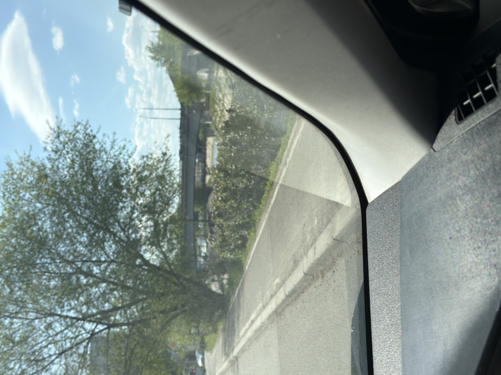 The photograph appears to be taken from inside a vehicle, looking out the driver's side window. The window frames a view of a road with a white line marking the edge. A large tree with green leaves is visible on the left side of the frame, and the sky is blue with scattered clouds. Beyond the road, there's vegetation, possibly bushes or trees, and a structure that could be a bridge or overpass. The interior of the vehicle is visible on the right side of the frame, showing part of the dashboard and an air vent. The location is identified as Krasnoyarsk, Russia. The scene appears to be daytime, with natural light illuminating the landscape.