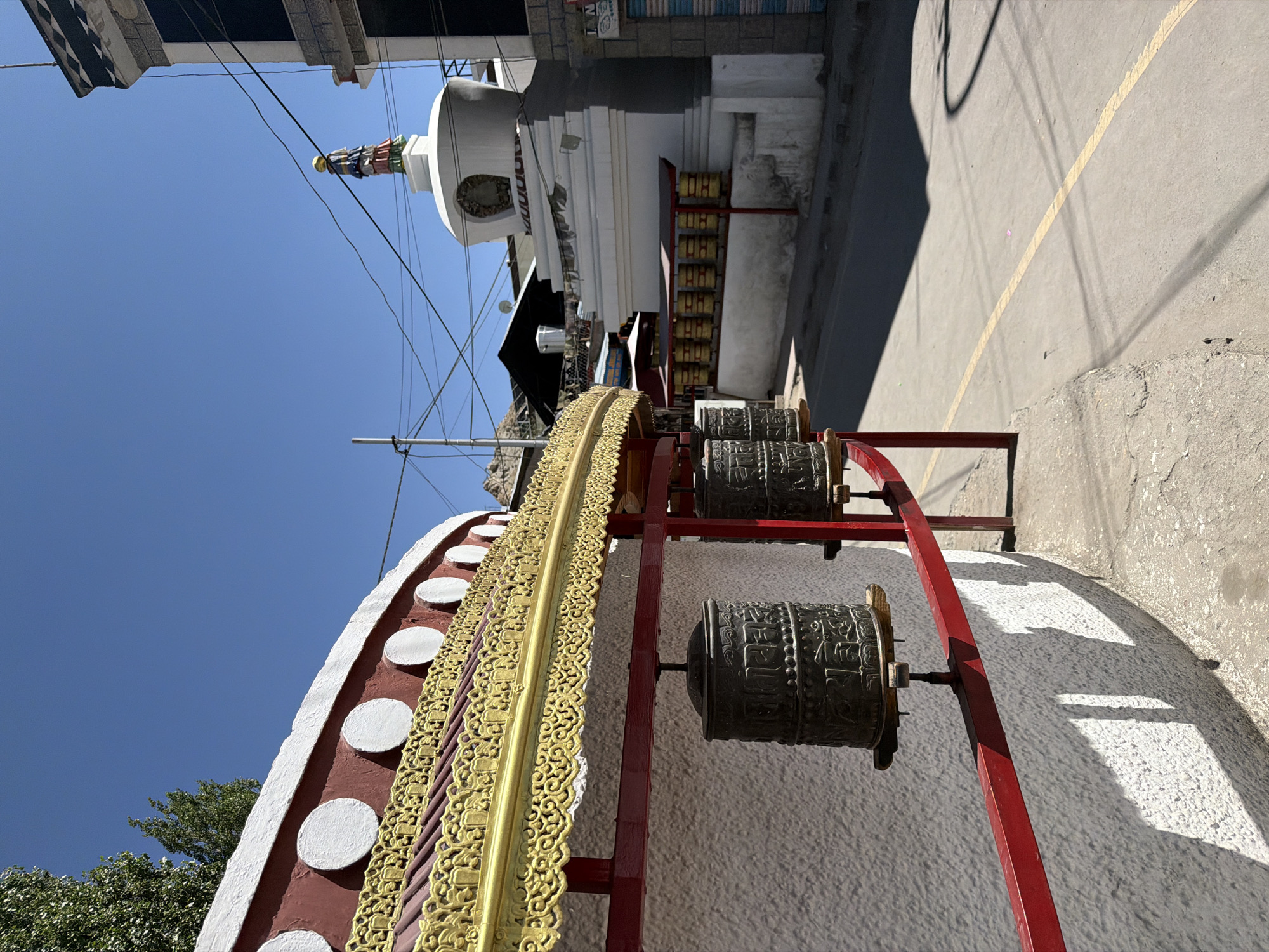This image captures a view in Leh, India, focusing on Buddhist elements and urban architecture under a clear blue sky. In the foreground, a white stucco wall is adorned with a decorative red and gold roofline, featuring circular white accents. Mounted on red metal poles are several dark, cylindrical prayer wheels, etched with intricate carvings. To the right, a white stupa, a Buddhist shrine, stands prominently. It is topped with a colorful statue and surrounded by a row of smaller, golden prayer wheels. Beyond the stupa, a modern building with dark windows and a patterned facade is visible, along with power lines crisscrossing the sky. The street in front of the stupa is paved with asphalt and marked with a yellow line. Trees and a hillside are visible in the background. The scene appears to be a street in Leh, likely near a monastery or temple complex.