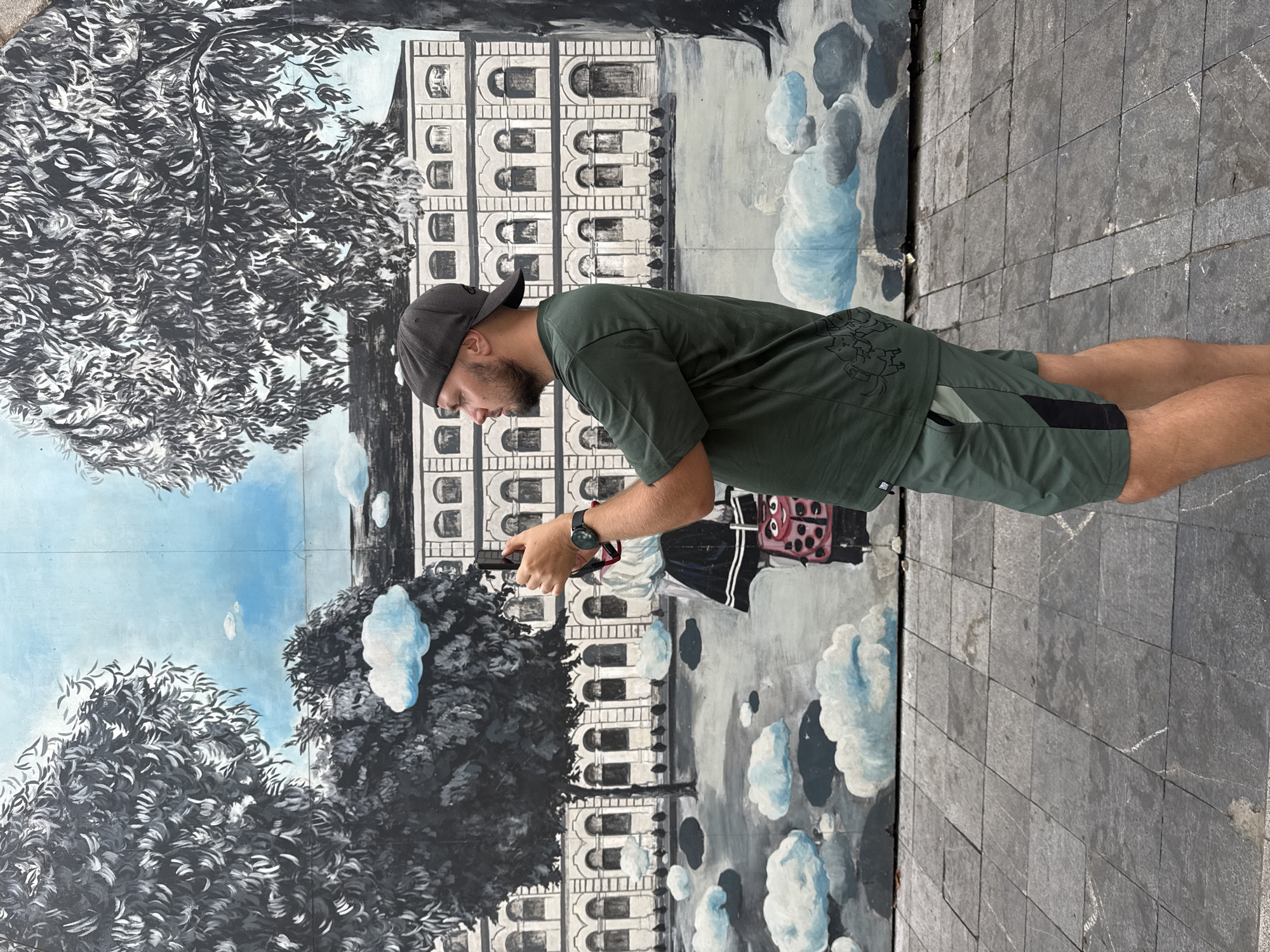 A man stands on a gray stone-tiled surface, possibly a sidewalk or plaza. He wears a dark green t-shirt, dark green shorts with a black stripe, a black baseball cap, and a wristwatch. He holds a smartphone in his hands, looking down at the screen. Behind the man is a painted mural on a wall. The mural depicts a building with multiple windows and an ornate facade, partially obscured by painted trees with black and white foliage. Also visible in the mural are painted clouds. The location appears to be in Hà Nội, Vietnam, based on the style of the mural and the types of stone used in the pavement.