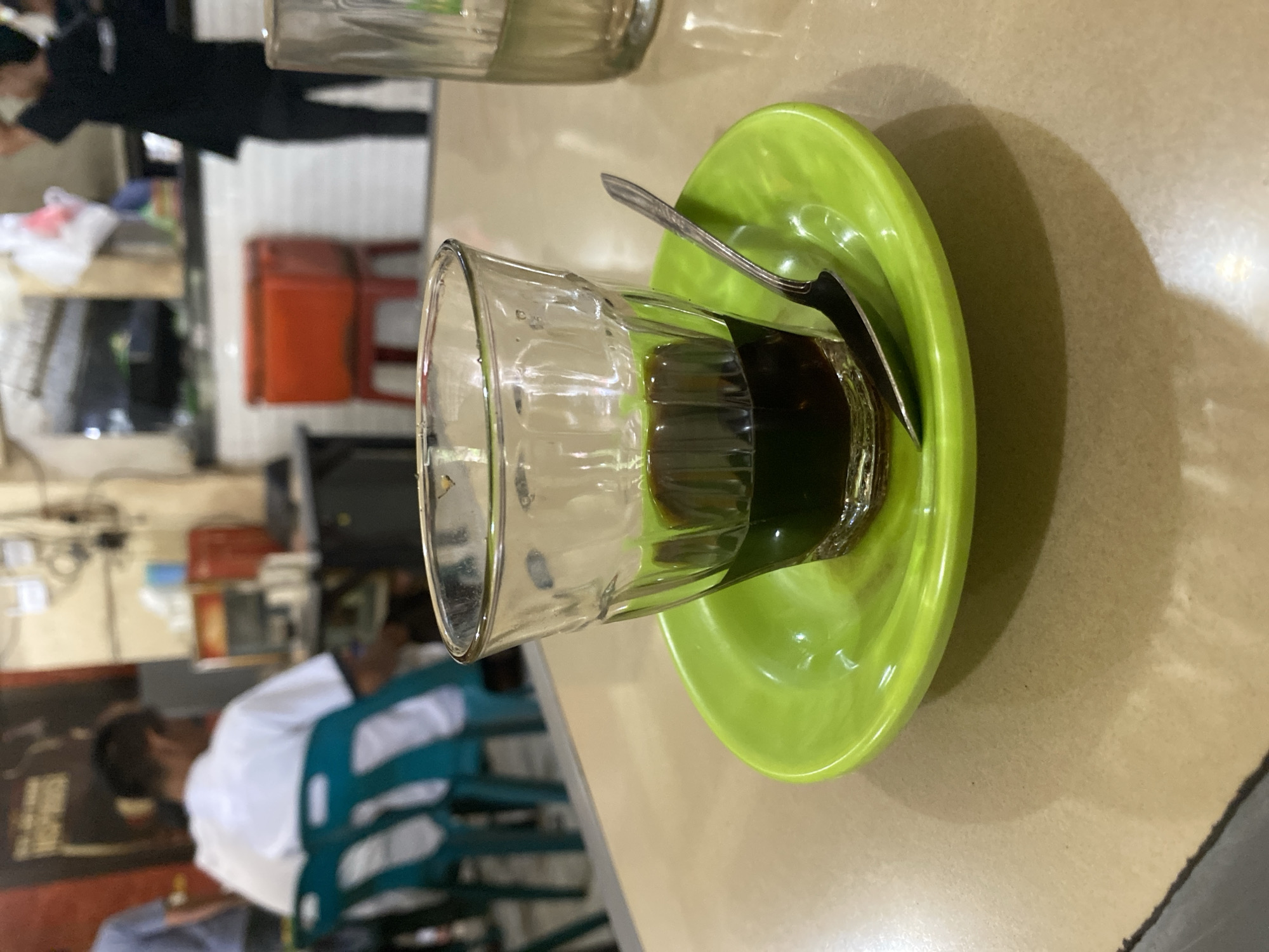 A close-up shot captures a glass of dark liquid, likely coffee, resting on a bright green saucer. A small, metallic spoon is partially submerged in the liquid and leans against the rim of the glass. The glass itself has a textured design. The scene appears to be set within a casual eatery or cafe, possibly in Lhokseumawe, Indonesia, given the context. In the blurred background, figures can be seen, including a person in a white shirt sitting and another individual in dark clothing standing near a counter. Elements like chairs, a red cooler, and shelving units are also visible, suggesting a lively, functional space. The lighting is somewhat dim, creating soft shadows on the table.