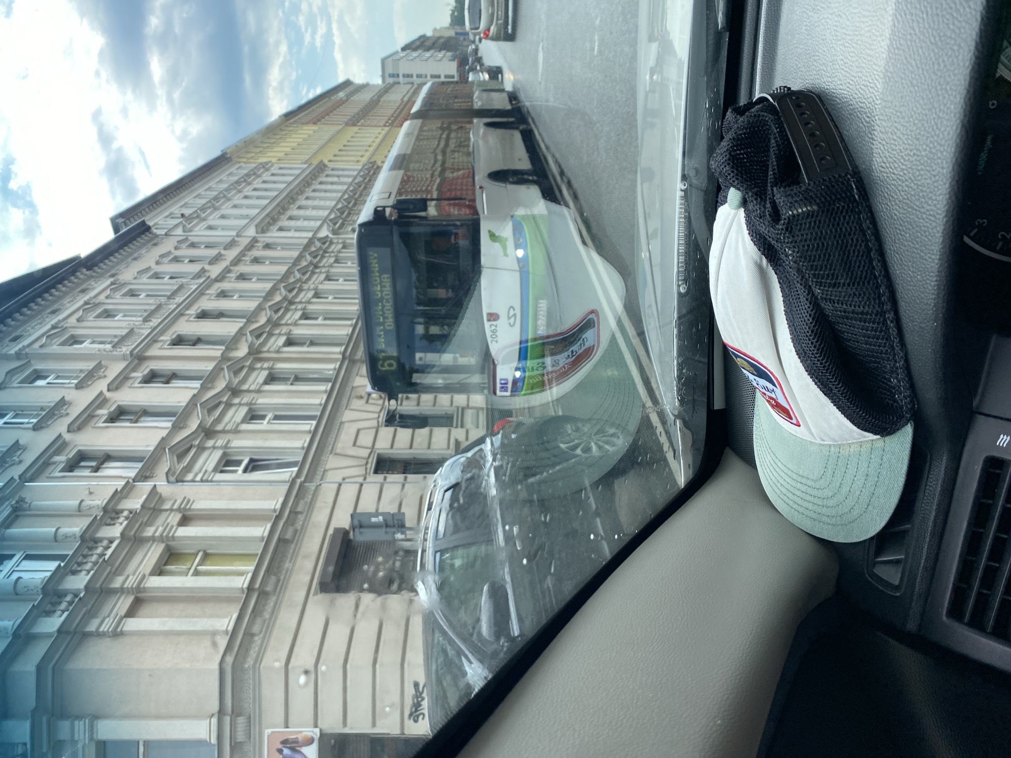 The image, taken from inside a vehicle, shows a street scene in Szczecin, Poland. The view is through the windshield, which is slightly wet, suggesting recent rain. The sky above is cloudy, indicating overcast weather.
The main subjects in the scene are a bus and the side of a building. The bus, likely a public transport vehicle, is white with green and blue accents, and displays route number 61. Text on the bus's destination board is partially visible and appears to be in a Slavic language, possibly Polish, with "61" clearly legible. The building is tall and multi-story, with a light-colored facade and numerous windows. Its architecture suggests an urban European setting.
Reflected on the windshield, a black mesh cap with a light blue brim and a red and white logo is visible, hanging from what appears to be a hook. This cap is an object within the vehicle and part of the foreground.
The image is captured from a low angle, looking upwards towards the sky and along the street. The perspective gives a sense of motion or being in transit. The overall mood is everyday and observational, without any strong emotional cues. The time of day is difficult to ascertain precisely due to the overcast conditions, but it appears to be daytime. The presence of the bus and buildings clearly places the scene in an urban environment, and the specific context of Szczecin, Poland, is provided.