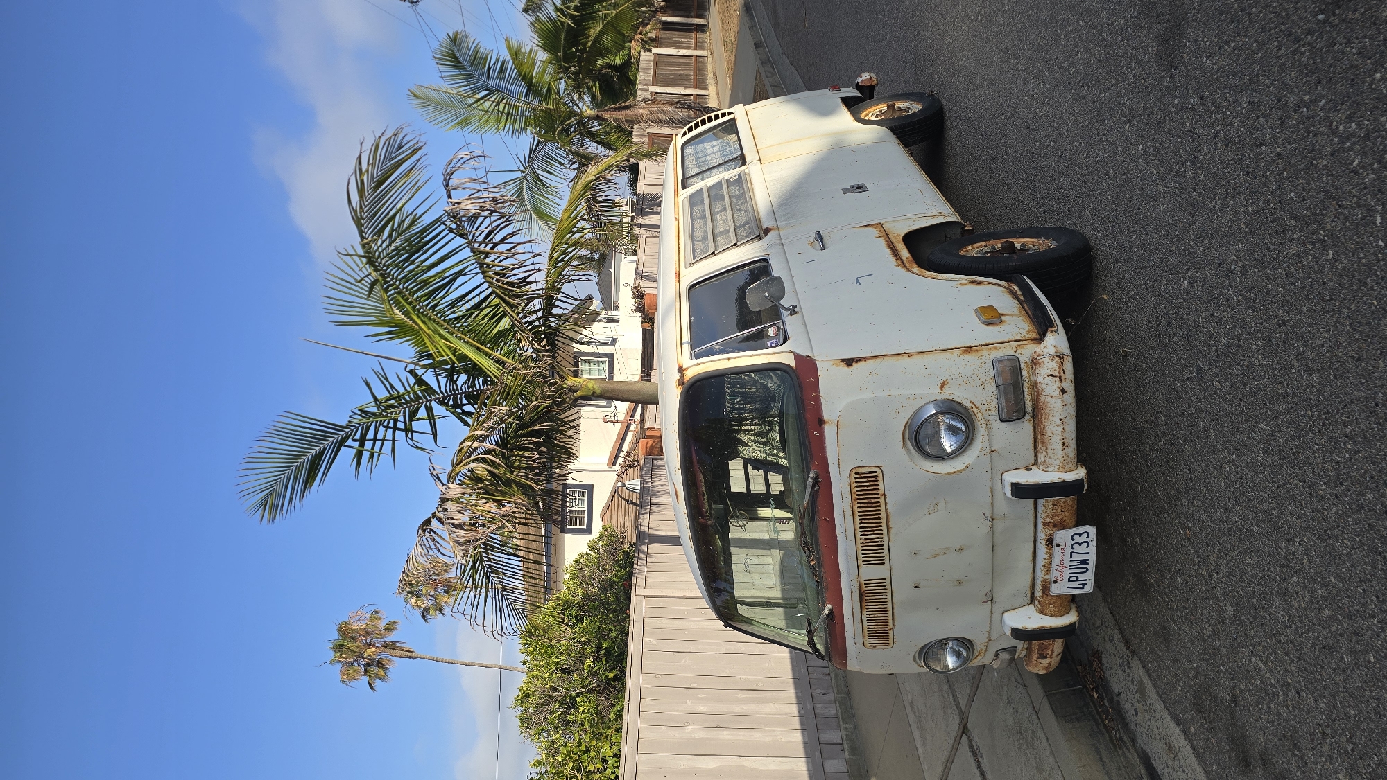 The image displays a weathered, white vintage van, likely a Volkswagen Type 2, parked parallel to a curb on a paved street. The van exhibits extensive rust along its body panels, particularly around the front bumper, headlights, and side seams. A California license plate, "4PUW733," is clearly visible on the front bumper.
The setting appears to be a residential street. To the right of the van, several large palm trees with green and some dried fronds dominate the midground, partially obscuring houses and a wooden fence. Another tall palm tree stands in the distance against the sky. The sky is bright blue with a few scattered white clouds, indicating a sunny daytime. There are no people visible, and no specific activity is taking place; the van is simply parked. The overall impression is that of a warm, coastal, or warm-climate environment.