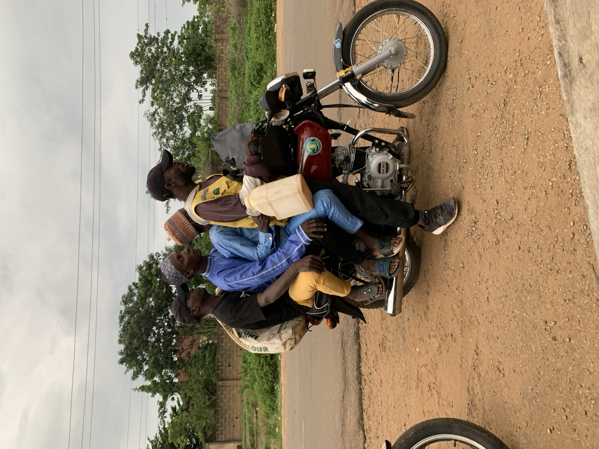 In Gbeleko, Nigeria, a side-profile view captures four males on a red Bajaj Boxer motorcycle, positioned on a reddish-brown dirt shoulder next to a paved road, under an overcast sky. The motorcycle appears stationary.
The driver, a dark-skinned male, wears a black baseball cap, a brown long-sleeved shirt, and a yellow sleeveless vest. A beige plastic jerrycan is suspended from the motorcycle's front, and a dark bag is draped over the handlebars.
Behind the driver, a dark-skinned male wears a blue long-sleeved tunic and a geometric-patterned white and black kufi.
The third passenger, appearing smaller, is largely obscured behind the second, wearing a light blue long-sleeved garment and a patterned kufi.
The rearmost passenger, a dark-skinned male, wears a black t-shirt and yellow pants. A large white sack, labeled "FLOUR" and "NIGERIA," rests on the motorcycle's rear, behind him.
All individuals wear sandals, except the driver who wears dark athletic shoes. The ground consists of dirt and small gravel. In the background, a low brick wall is partially visible, flanked by green trees and foliage. Overhead, power lines are present against the gray, cloudy sky. A portion of another motorcycle's front wheel is visible on the far left.