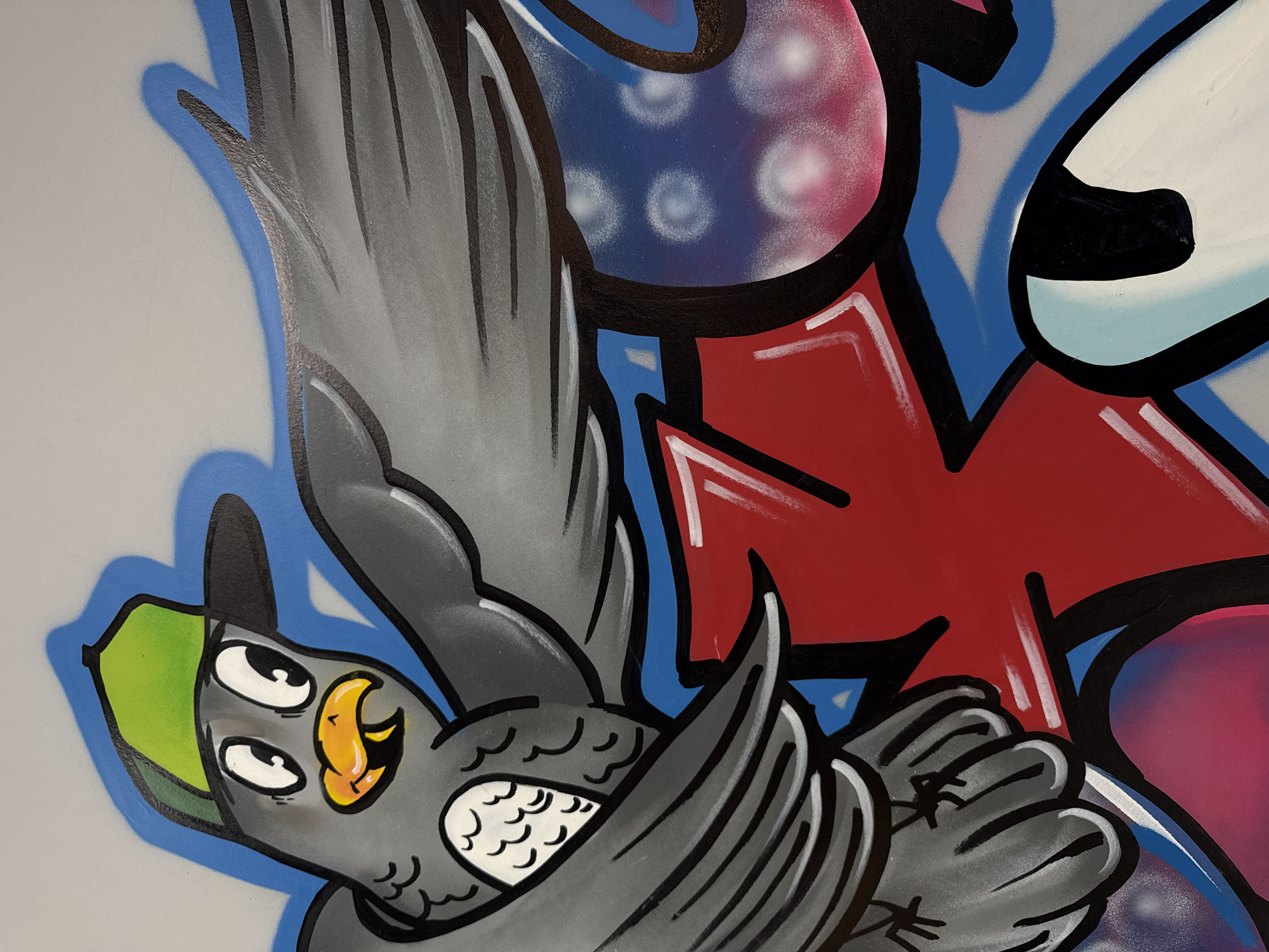 This media file captures a close-up section of a vibrant graffiti mural, located in Vladivostok, Russia. The artwork features a dynamic, cartoonish bird-like character prominently, set against a backdrop of bold, abstract shapes and stylized elements.
The central subject is a gray bird, depicted in a spirited, perhaps flying, pose with its wings spread. It possesses striking features, including two large, white, wide-open eyes with black pupils and a distinctive yellow and orange beak. The bird wears a green cap with a dark brim, adding to its character. Its body and wings are detailed with various shades of gray, emphasized by thick black outlines and white highlights, particularly on the feathers. A lighter blue spray-painted outline around the bird creates a shadow effect, making it stand out from the background.
Surrounding and intertwined with the bird are large, brightly colored graffiti elements. These include a significant red shape marked with white streaks, a blue section adorned with scattered white circular patterns resembling spray paint dots, and a pink or magenta area featuring similar textural details. A white shape with a light blue shadow is also visible. All these elements are sharply defined by thick black outlines. Broader strokes of blue paint are visible in the background, adding depth.
The overall aesthetic is energetic and expressive, typical of street art, characterized by its bold lines, contrasting colors, and a playful, lively mood. While the abstract forms hint at stylized lettering, no explicit or readable text is visible in the frame.